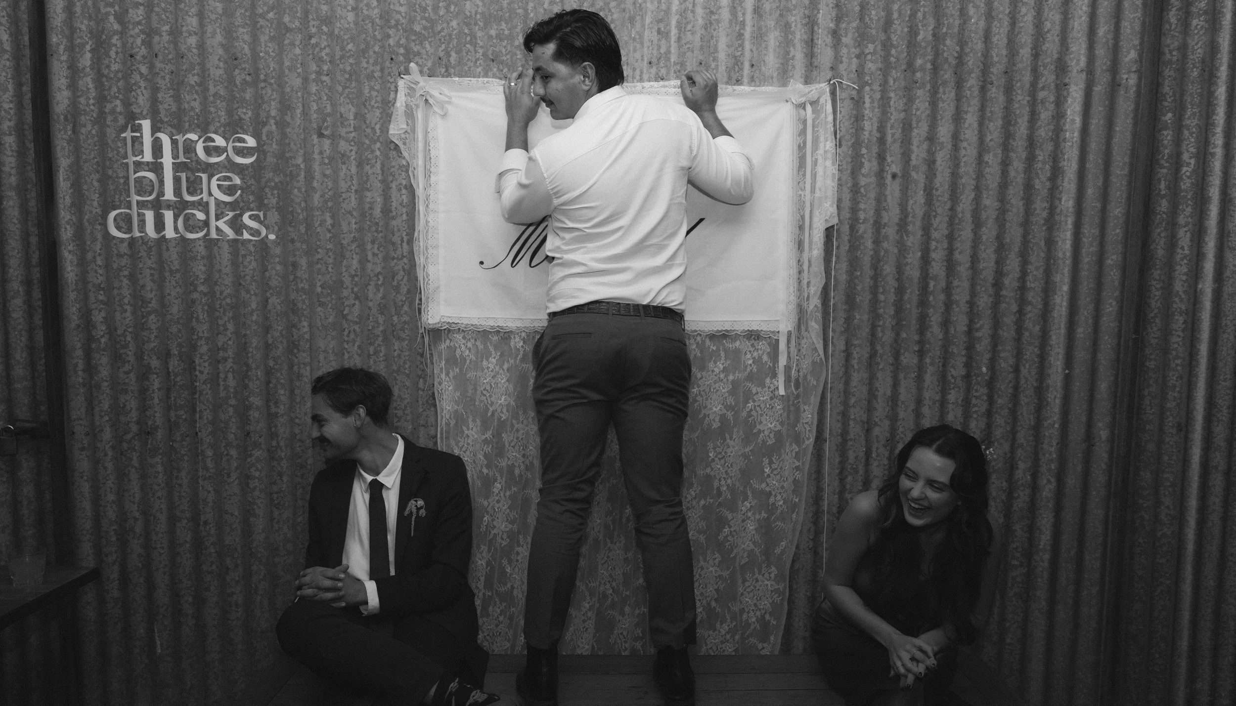 Black and white photo of three people at a party or event, with the man standing in the center with his back to the camera. He is adjusting a sign on a wall that says 'M' and part of another letter, surrounded by lace and fabric decoration. The man is wearing a light shirt and dark pants, sitting on the floor to the left is a man in a suit looking down, and to the right is a woman in dark clothing smiling and looking at the man in the center. There is a sign on the wall that reads 'three blue ducks.'