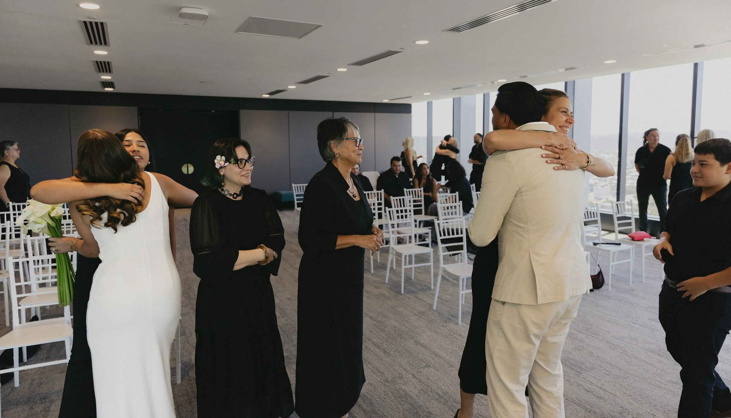 Groom hugging bride during a wedding ceremony with guests and friends in a modern venue with large windows.