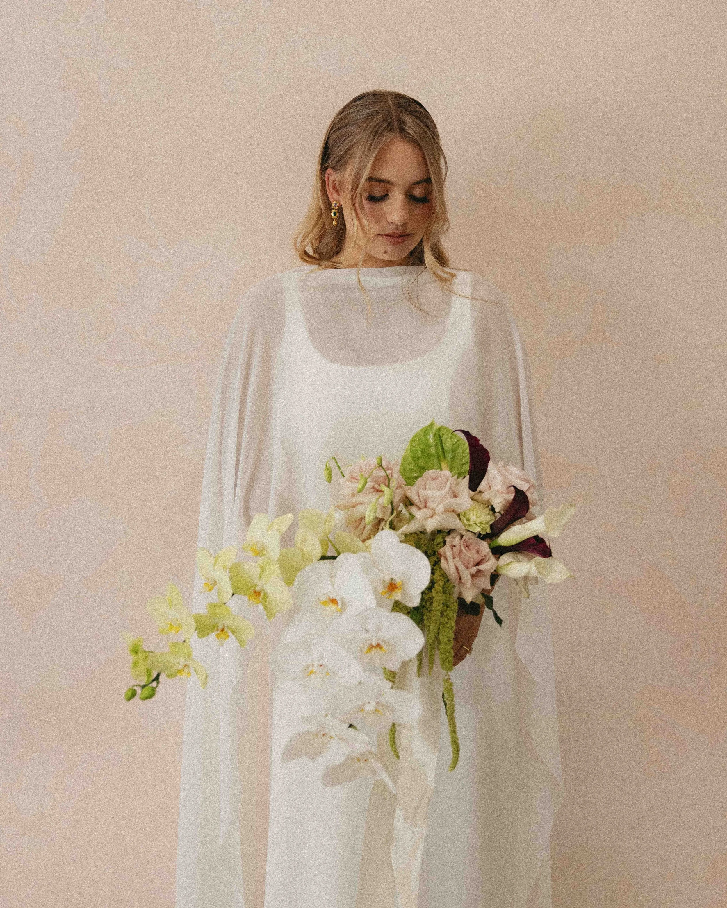 A woman in white dress holding a bouquet of roses and orchids against a beige background.
