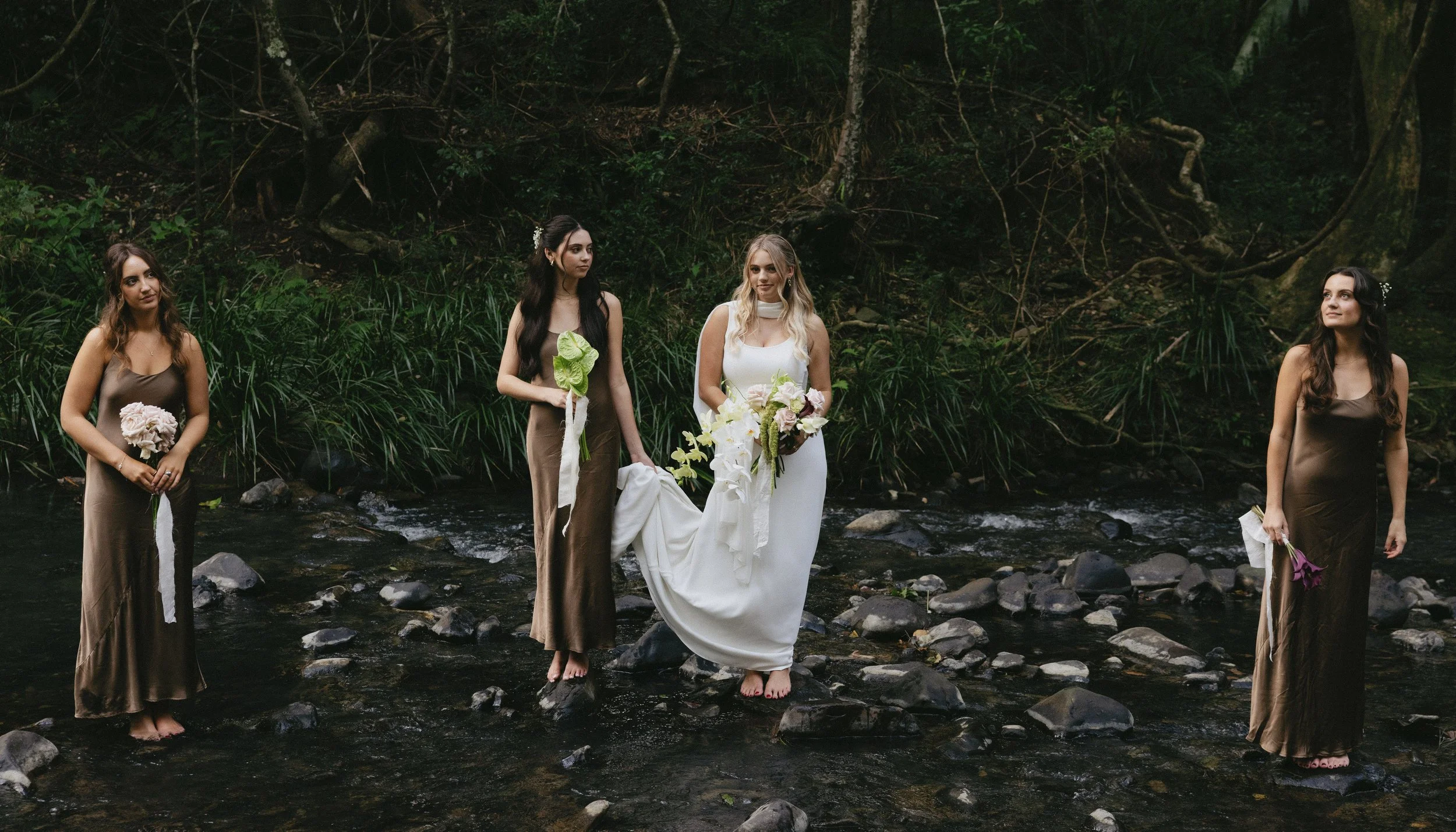 A group of five women standing in a shallow creek surrounded by greenery and trees, participating in a wedding or spiritual ceremony.