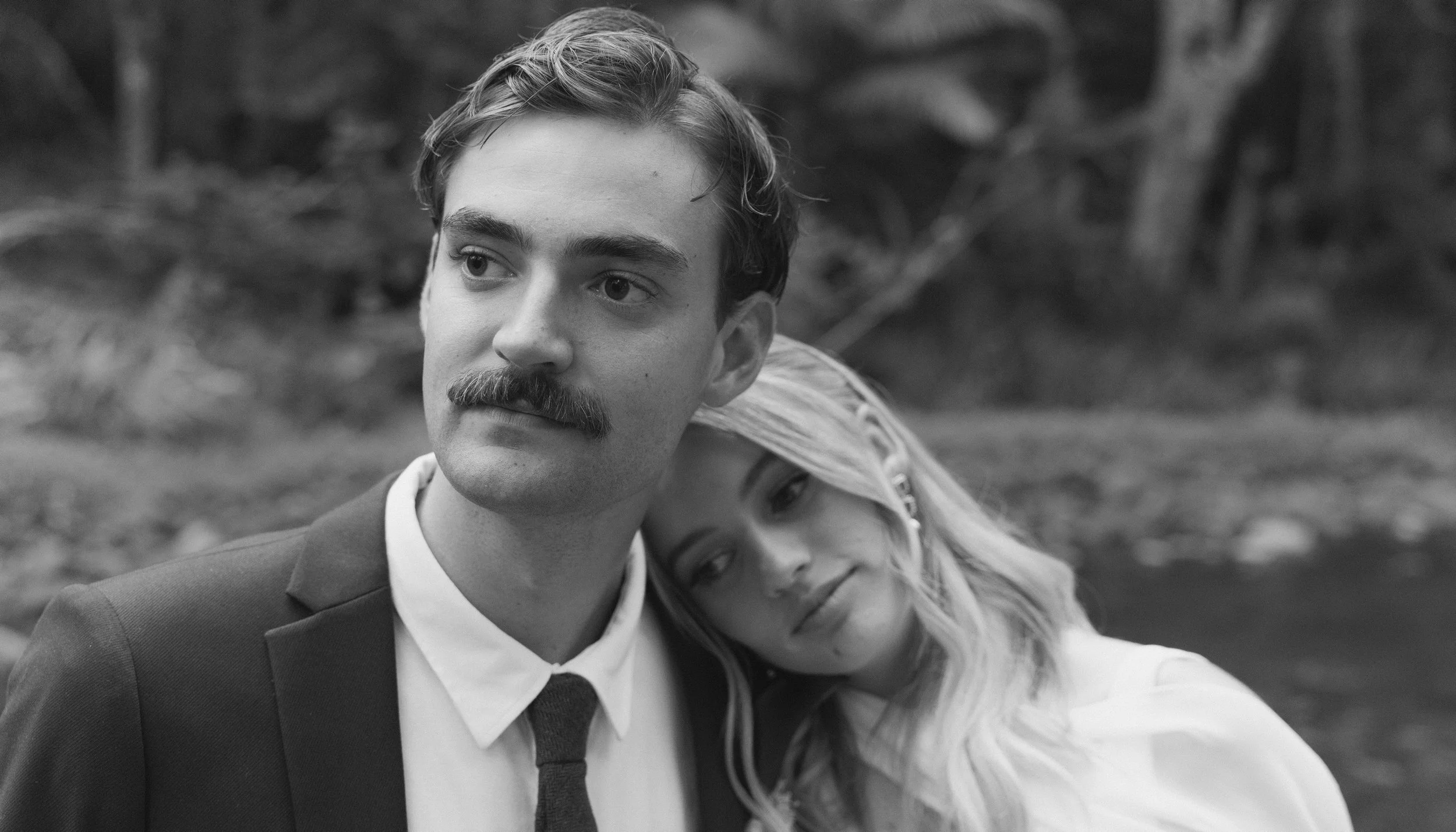 Black and white photo of a man with a mustache and suit, and a woman with long hair, resting her head on his shoulder, outdoors.