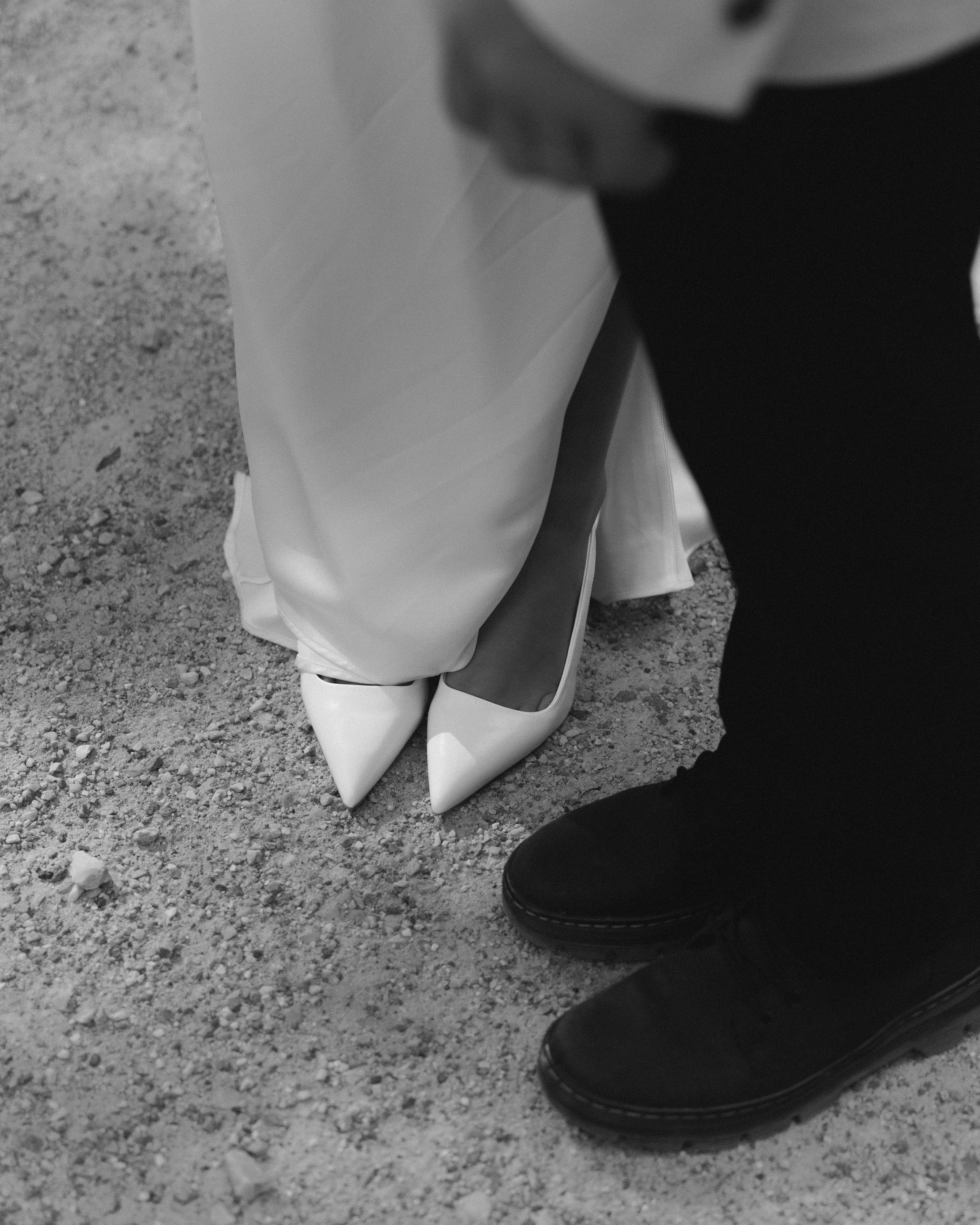 Close-up of a person in white pointed high heels and a long dress, with a person in black shoes assisting or adjusting the shoe on a gravel surface.