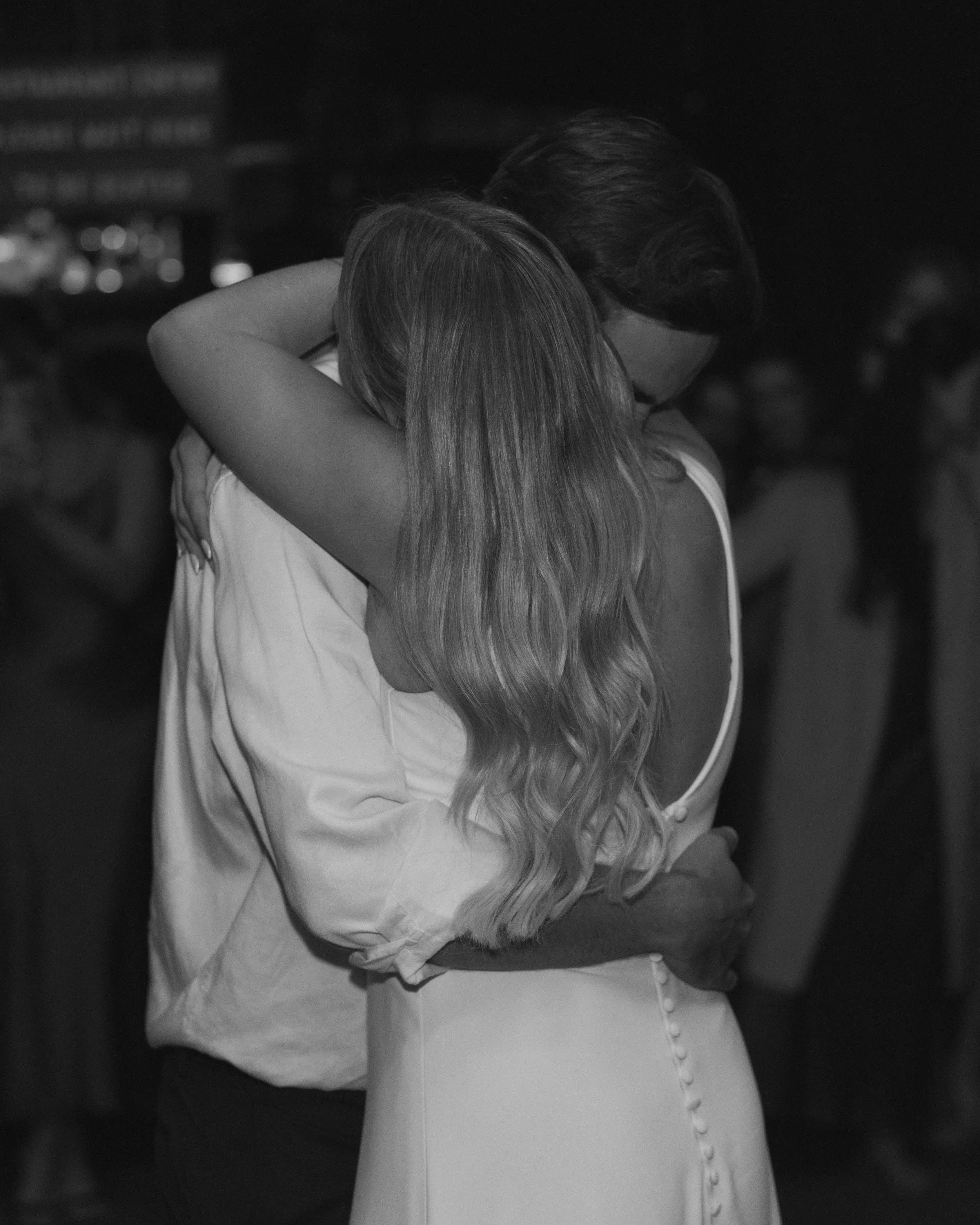A black-and-white photo of a couple embracing, with the woman’s long, wavy hair and the man’s face partially obscured, in a nighttime setting with blurred background.