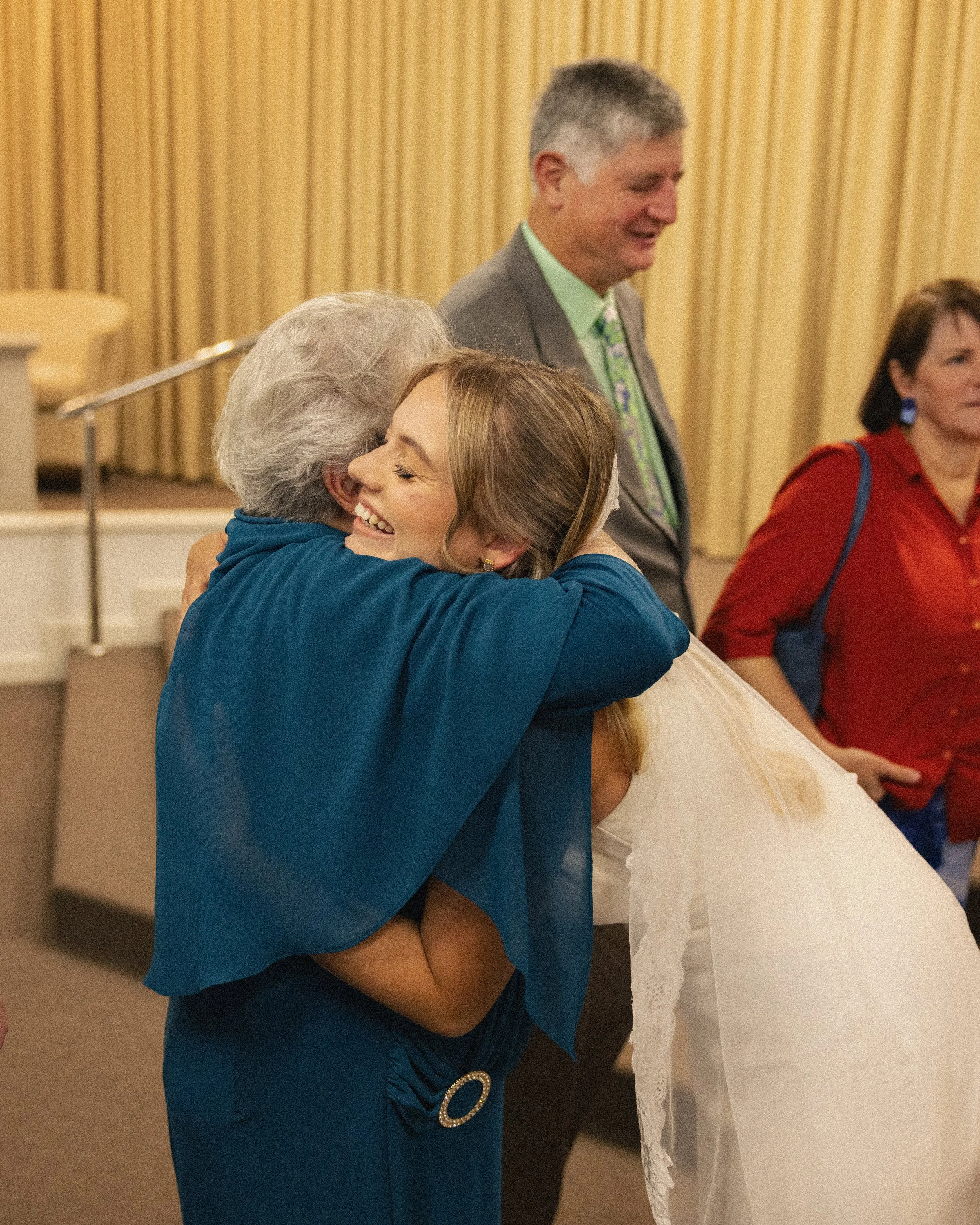 A woman in a blue dress hugging an elderly woman in white dress, smiling and embracing warmly, in a formal event setting with other people in the background.