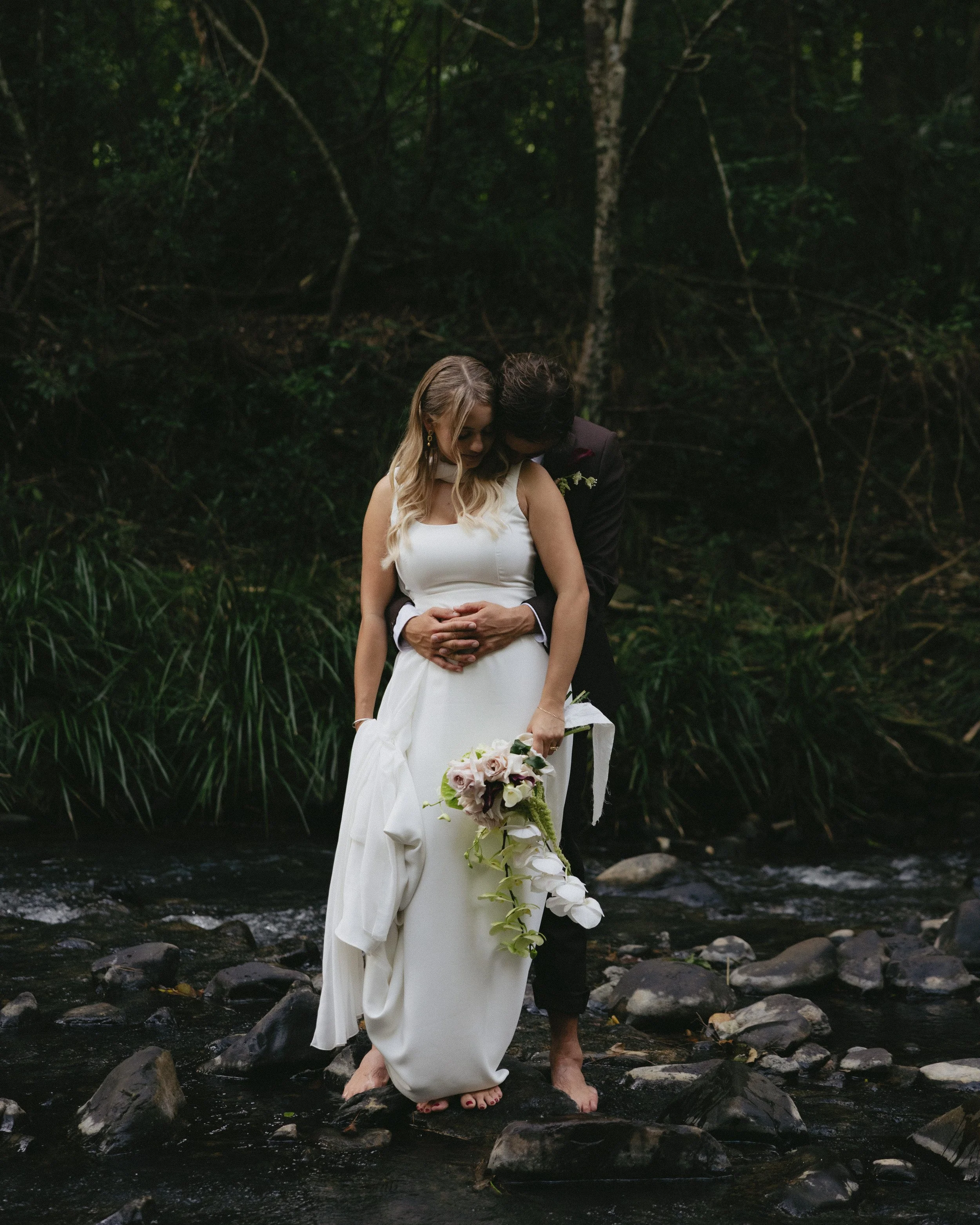 A bride and groom standing in a shallow stream in a forest, embracing each other. The bride holds a bouquet of flowers and wears a white dress, while the groom is dressed in a dark suit.