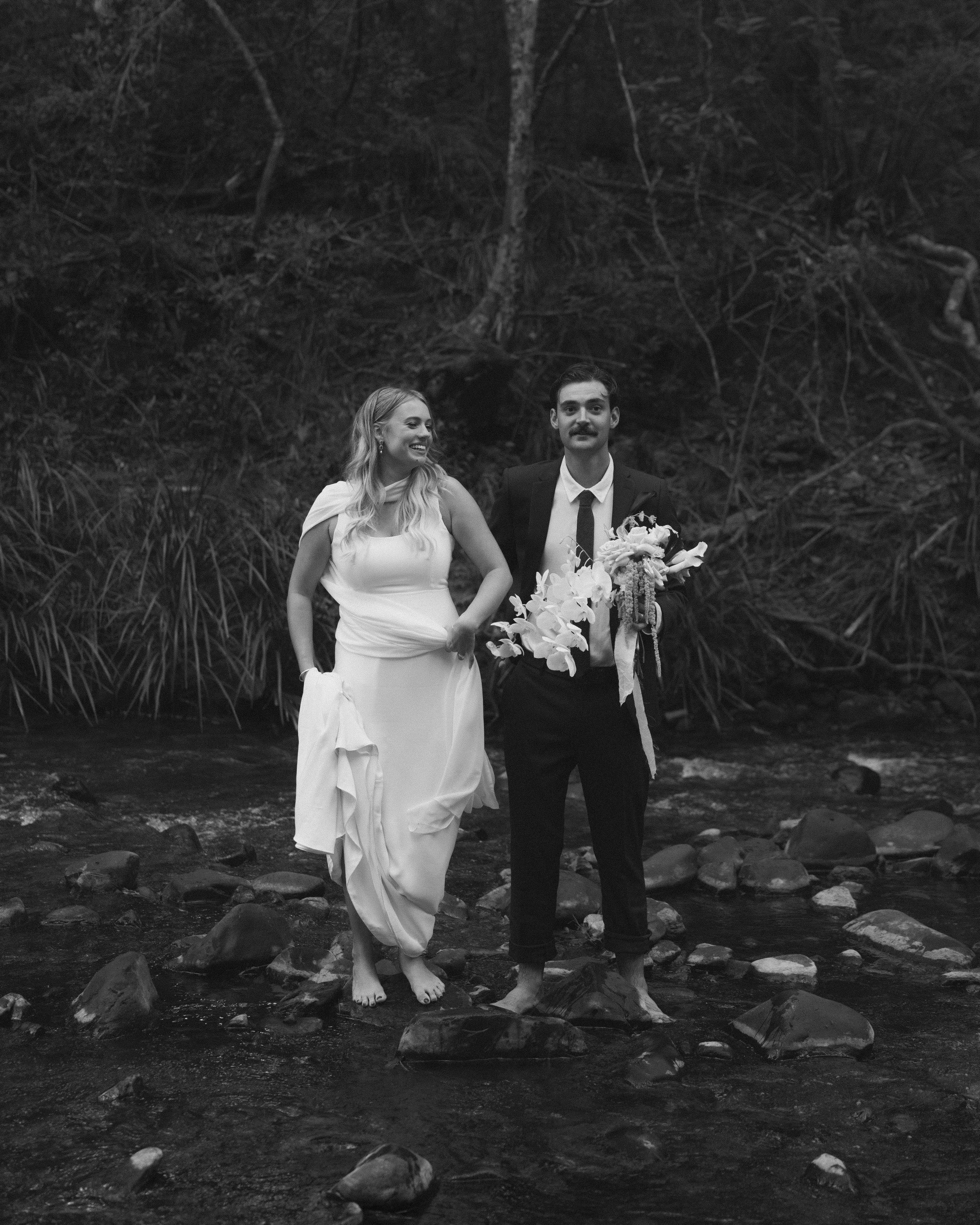 Black and white photo of a couple standing in a rocky stream surrounded by trees; the woman wears a white dress and the man a suit, holding a bouquet of flowers.