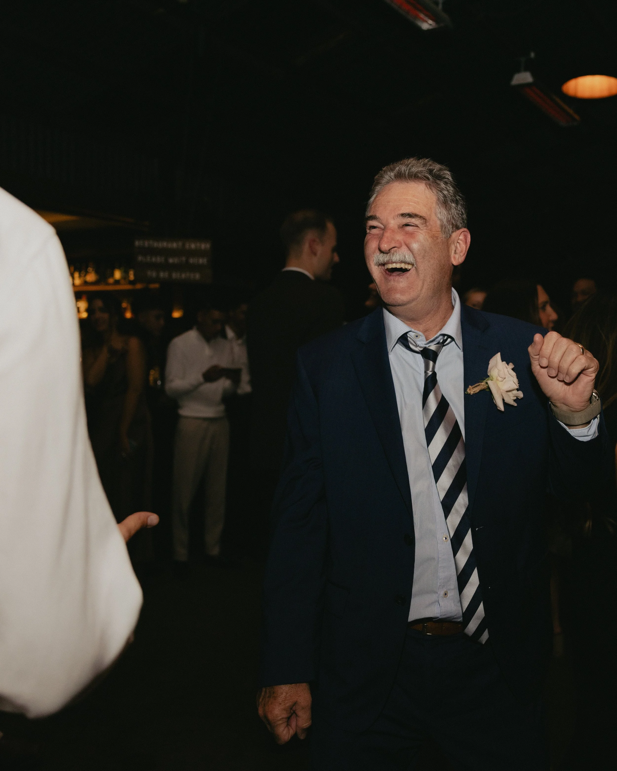 A man in a dark blue suit, striped tie, and white dress shirt, smiling and celebrating at a social event or party.