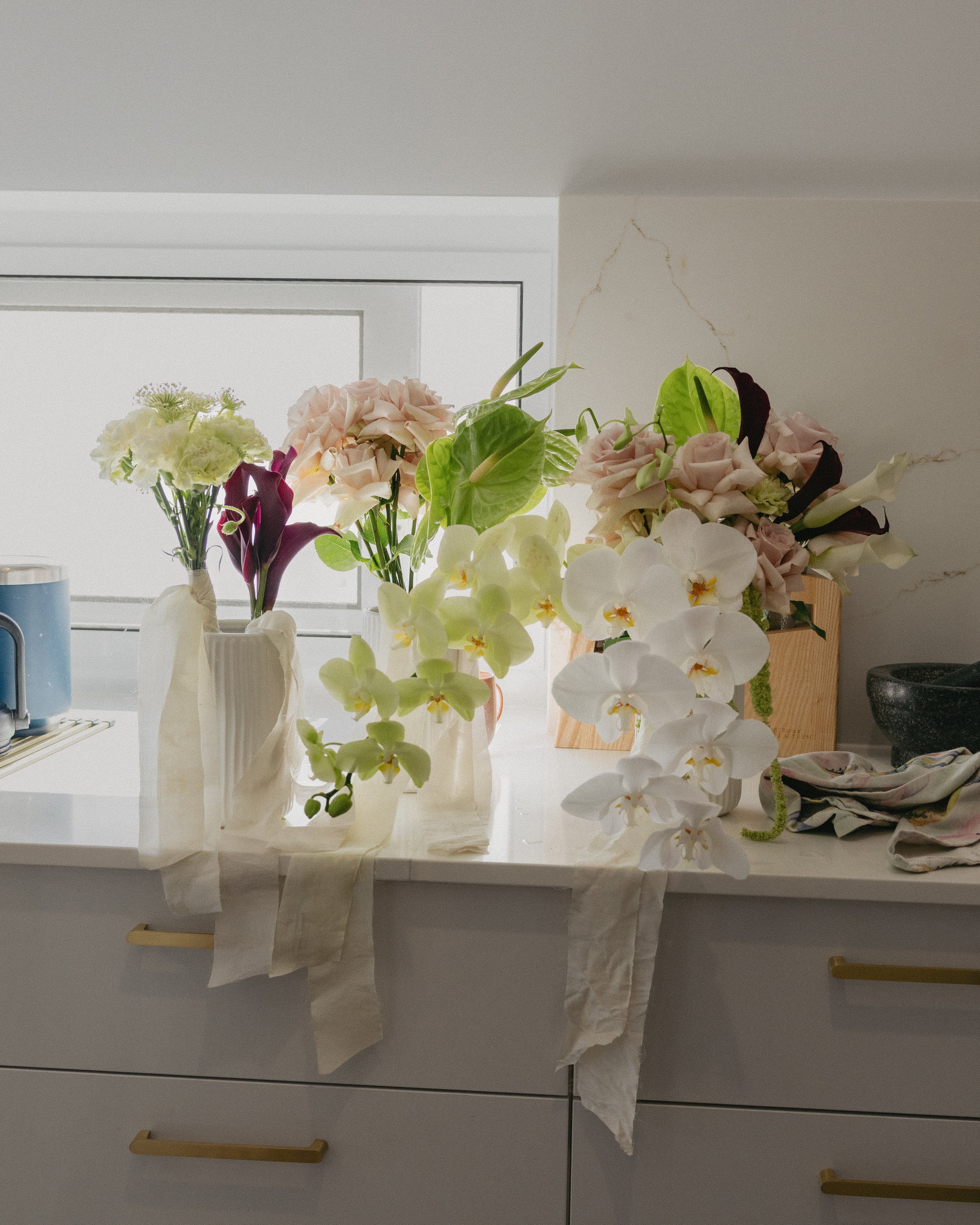 Arranged flowers in vases on a kitchen counter near a window, including orchids, calla lilies, and other blossoms with fabric and wooden decor.