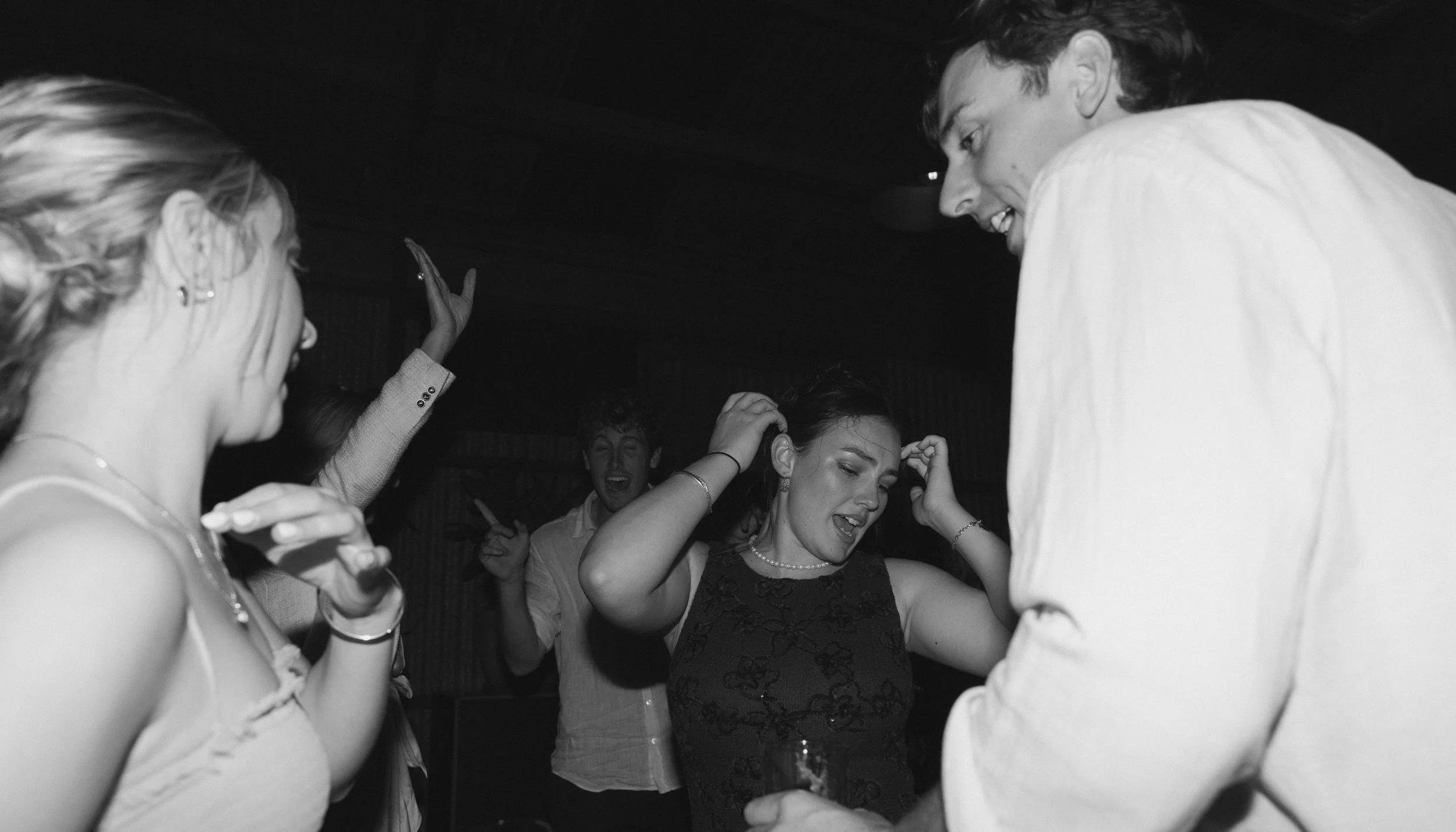 People dancing and having fun at a party, with a woman in the center adjusting her hair and a man on the right smiling at her, in black and white.