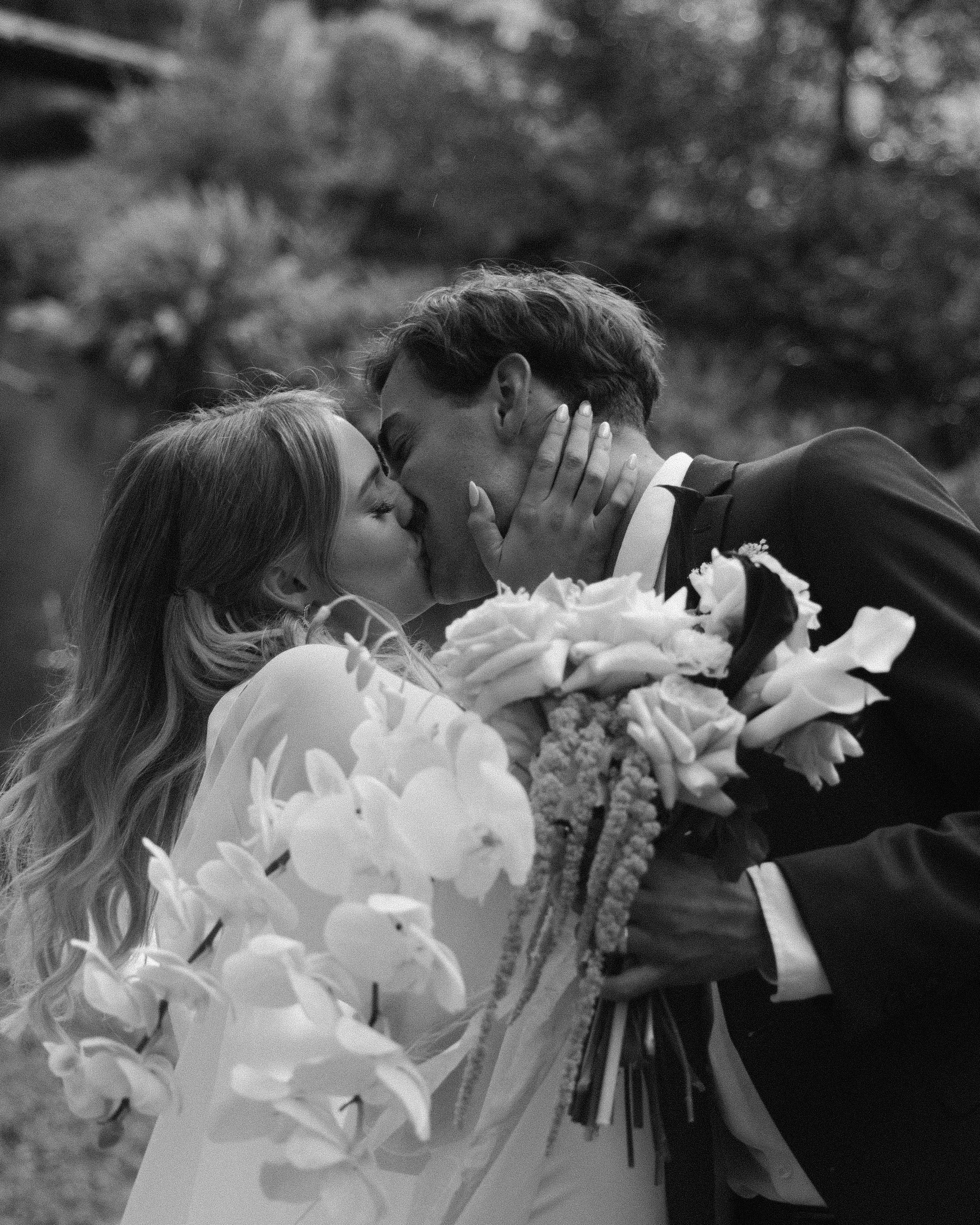 A black and white photo of a couple kissing with a large bouquet of flowers, outdoors with blurred trees in the background.
