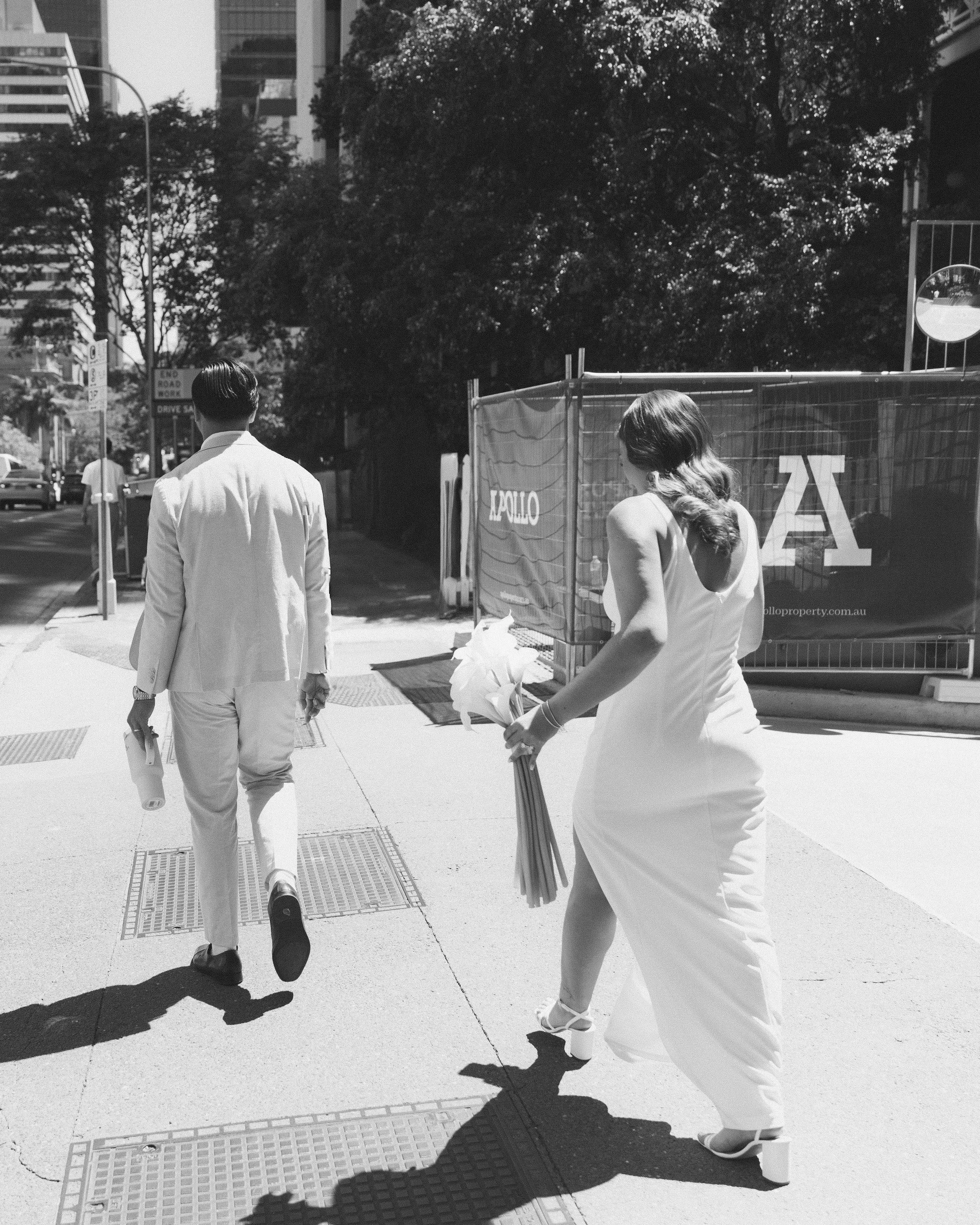 A bride in a white wedding dress holding a bouquet of flowers walking on a city sidewalk with a groom in a white suit, both walking away from the camera on a sunny day.
