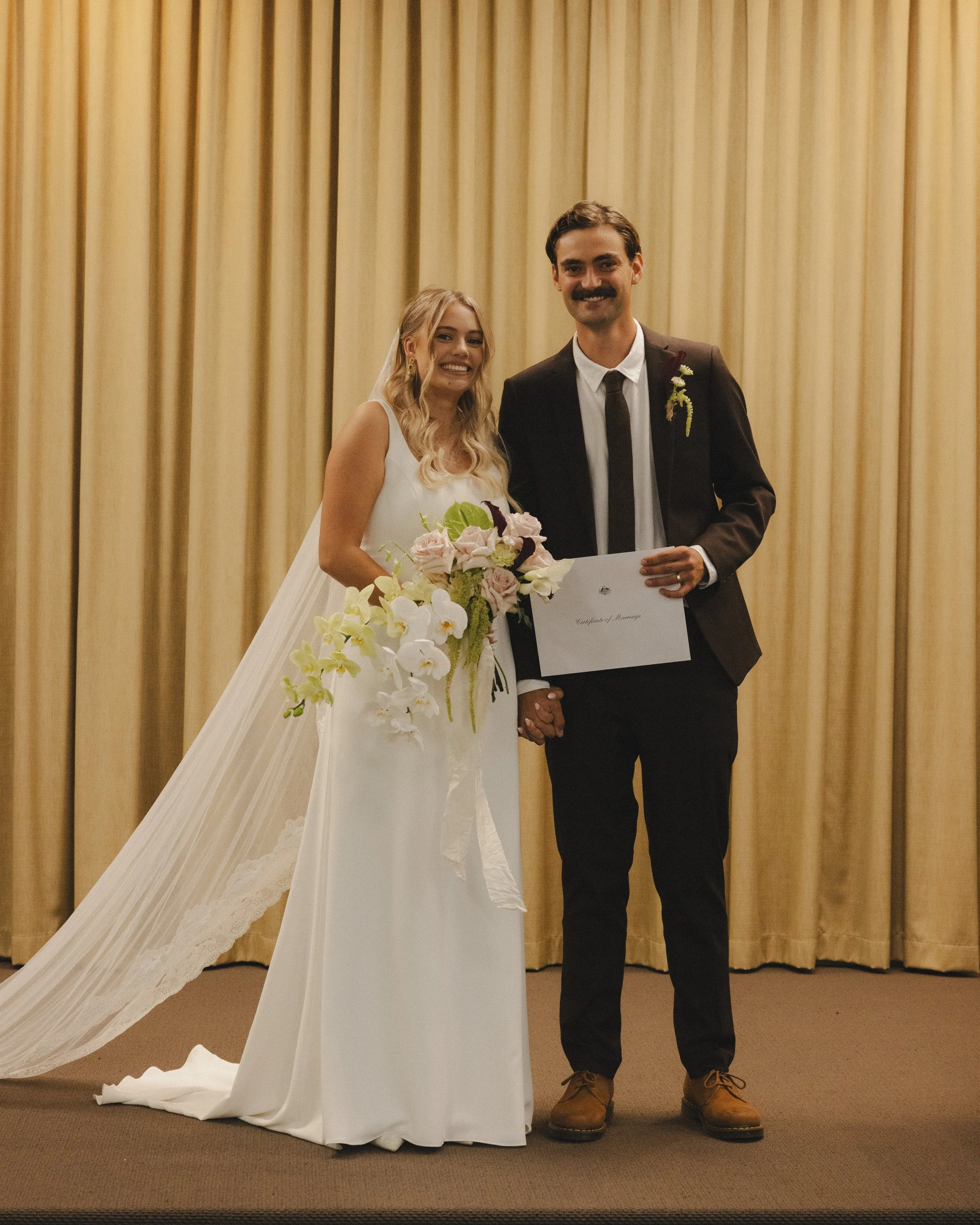A bride and groom standing together holding hands, smiling, with the groom holding a marriage certificate, against a gold curtain backdrop.