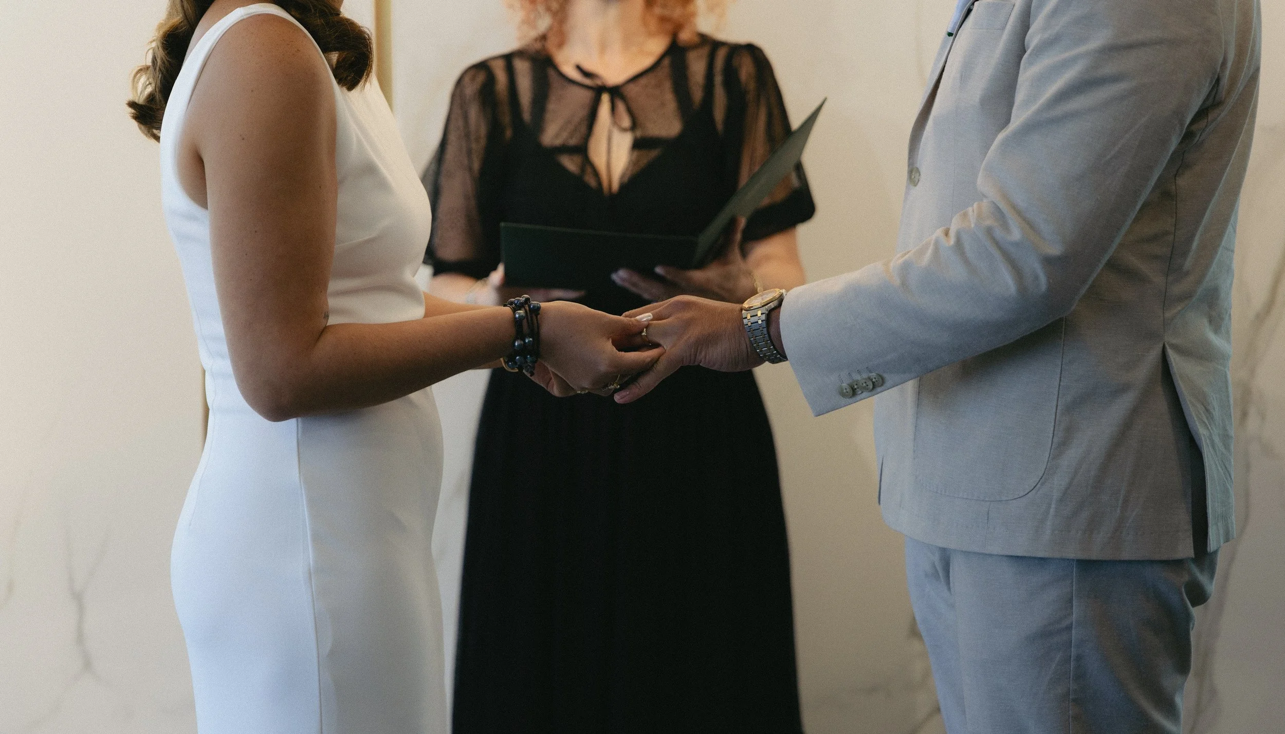 A couple holding hands during their wedding ceremony, with an officiant reading from a black folder in the background.