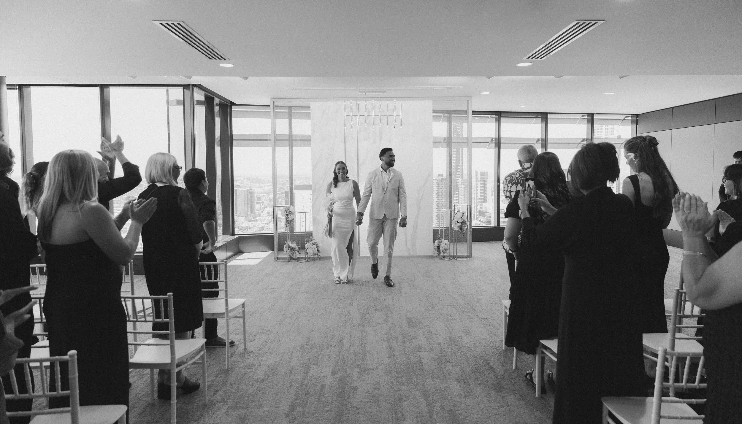 A black-and-white photo of a wedding ceremony inside a modern high-rise building with large windows showing a city skyline. The bride and groom are walking down the aisle, holding hands, while guests are standing and clapping, some taking photos.