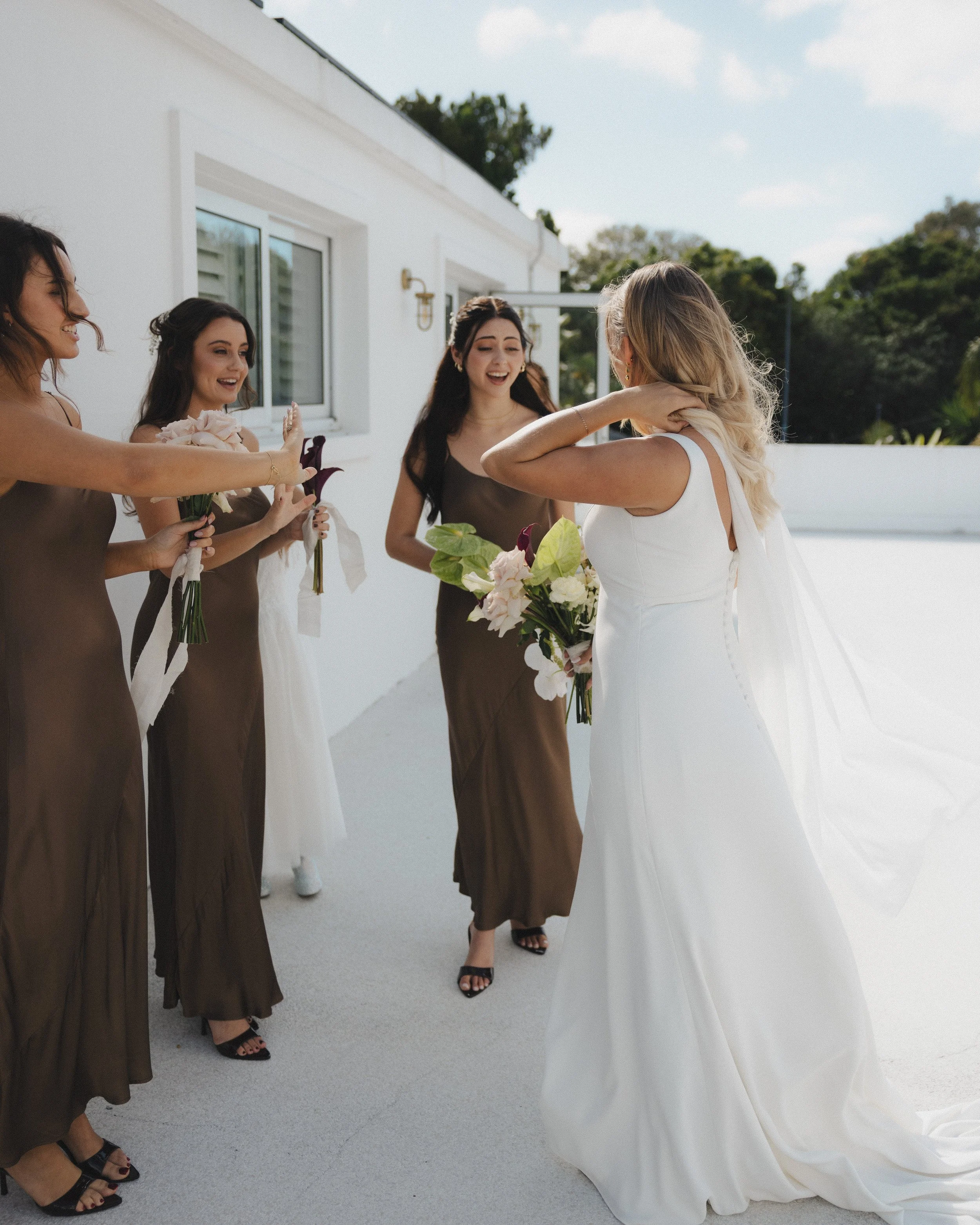 A bride with long blonde hair in a white wedding dress is smiling and holding a bouquet of flowers, standing in front of four bridesmaids who are dressed in brown gowns, on a terrace outdoors with trees and a building in the background.