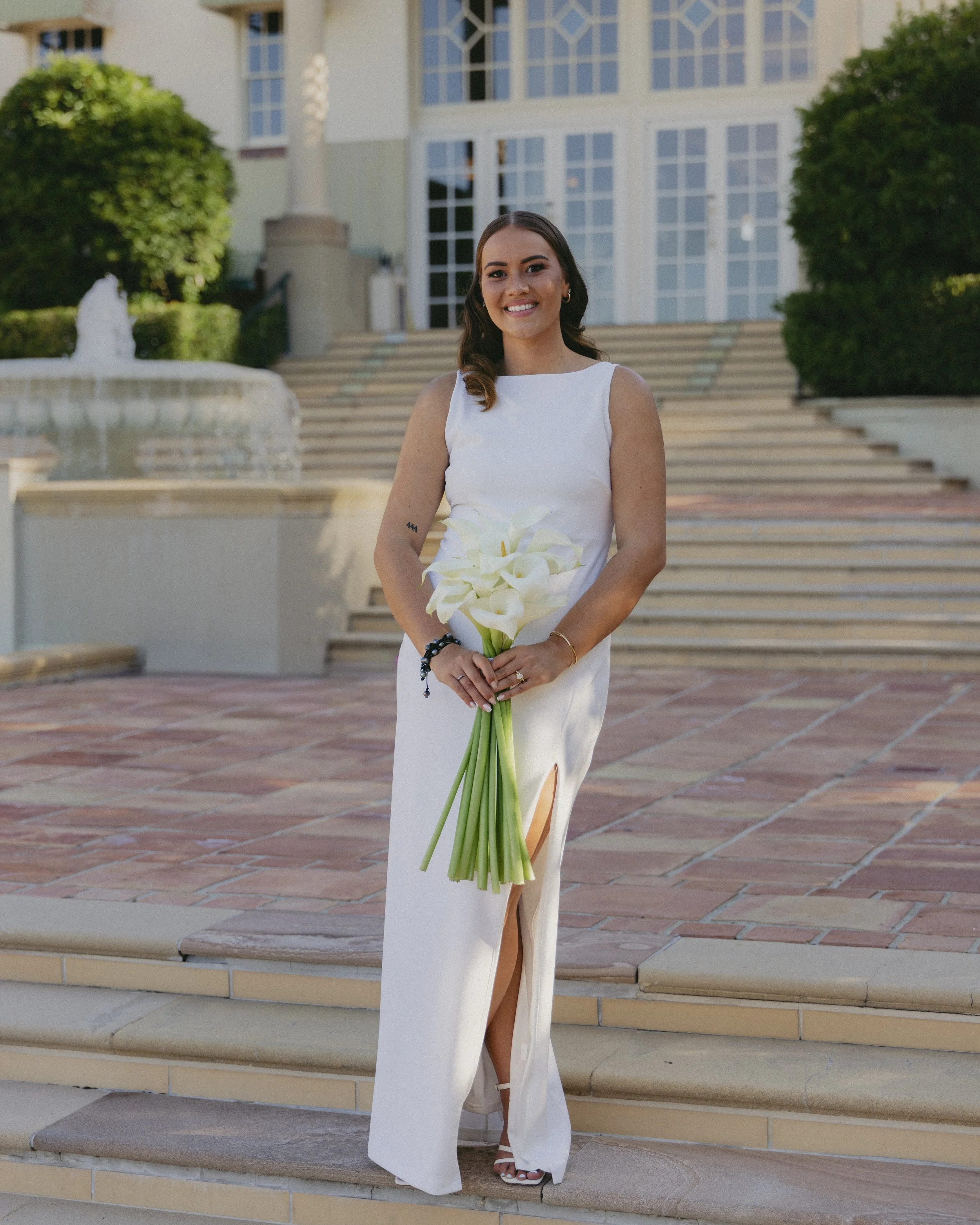 A woman in a white dress standing outdoors on steps, holding a bouquet of calla lilies, smiling at the camera.