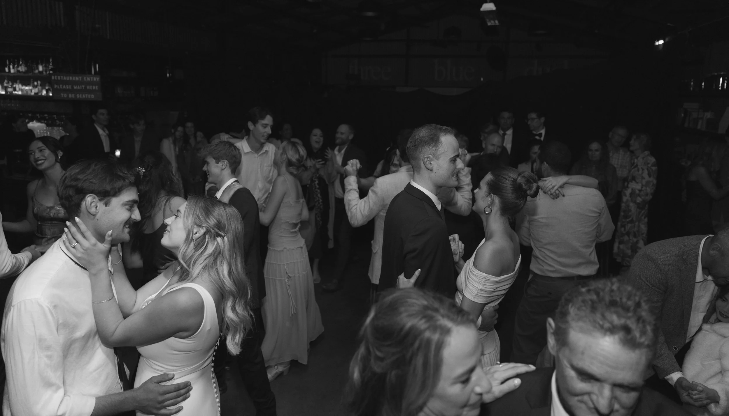 People dancing closely together at a lively party or celebration, with some couples gazing into each other's eyes, in a dimly lit venue.