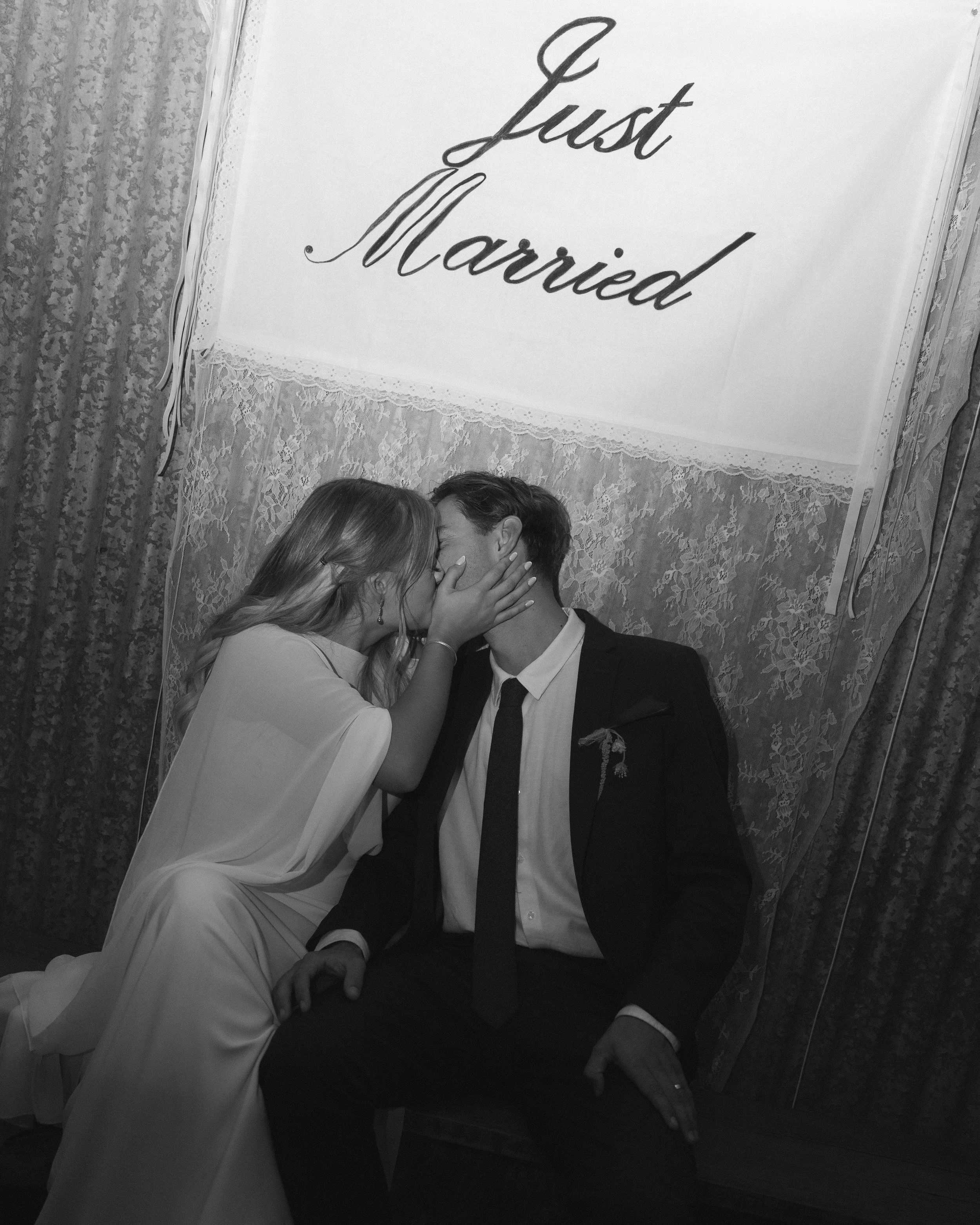 A bride and groom kissing in front of a sign that says 'Just Married' at their wedding reception, black and white photo.