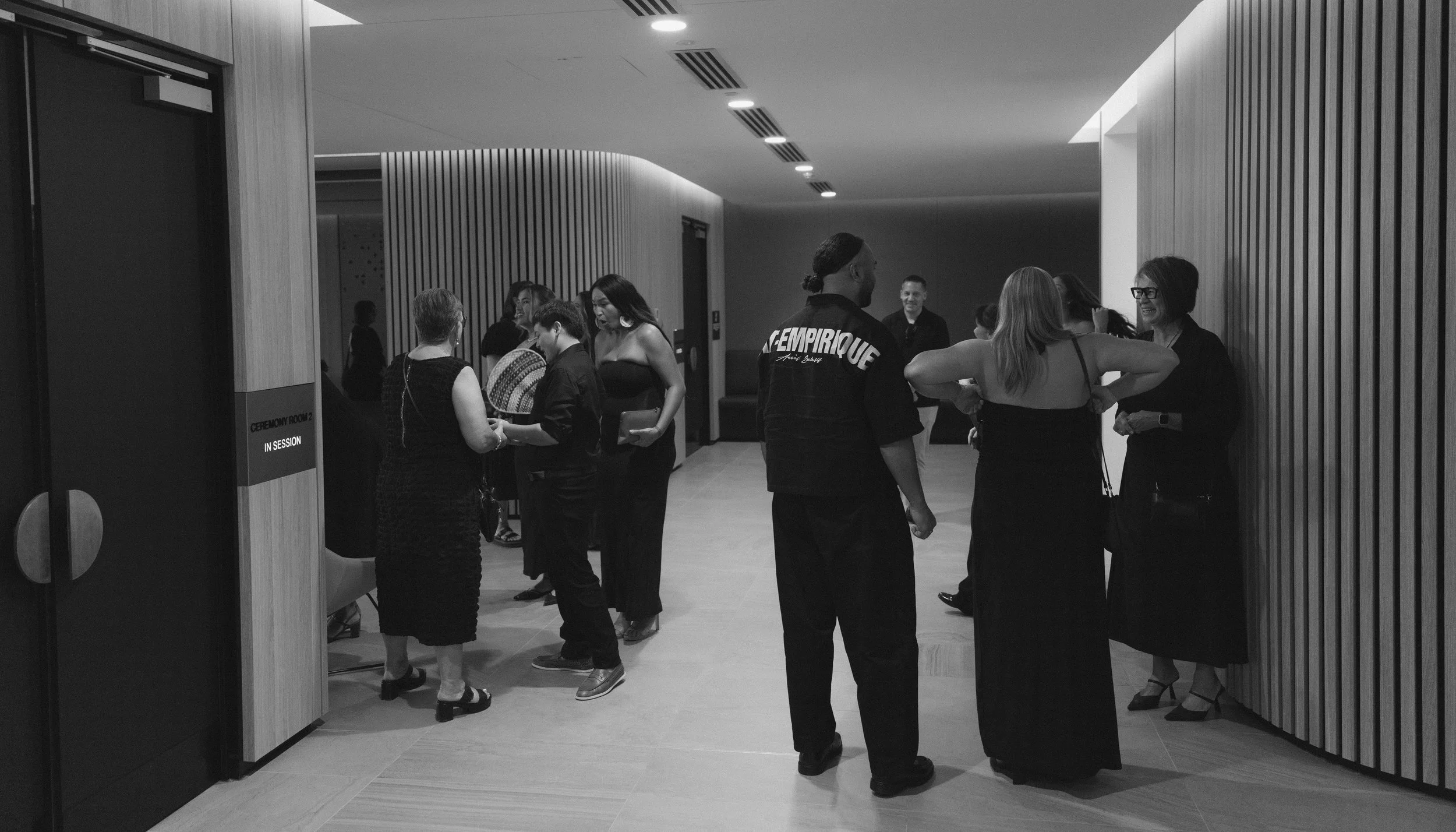 Group of people socializing in a modern interior space, standing and talking in small groups, with wood panel walls and soft lighting.