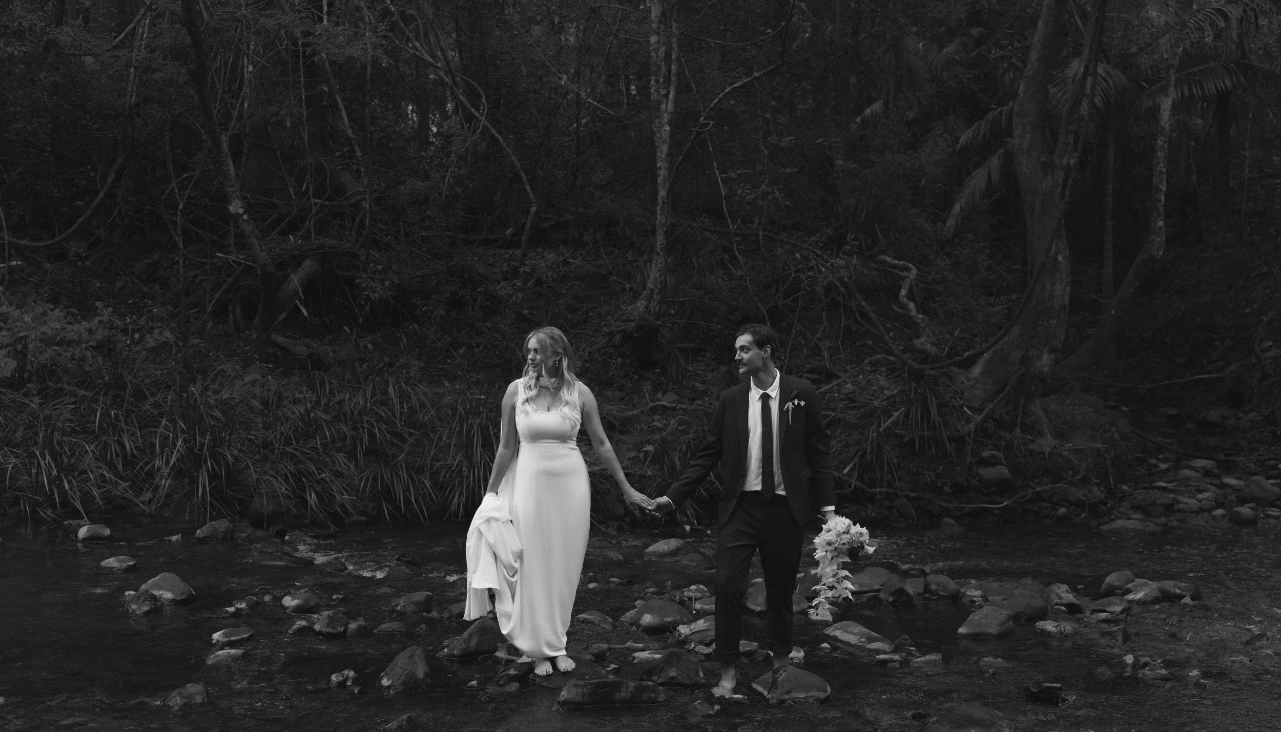 A black and white photo of a bride and groom holding hands and walking through a shallow creek in a forested area, with the bride wearing a long white dress and the groom in a suit.