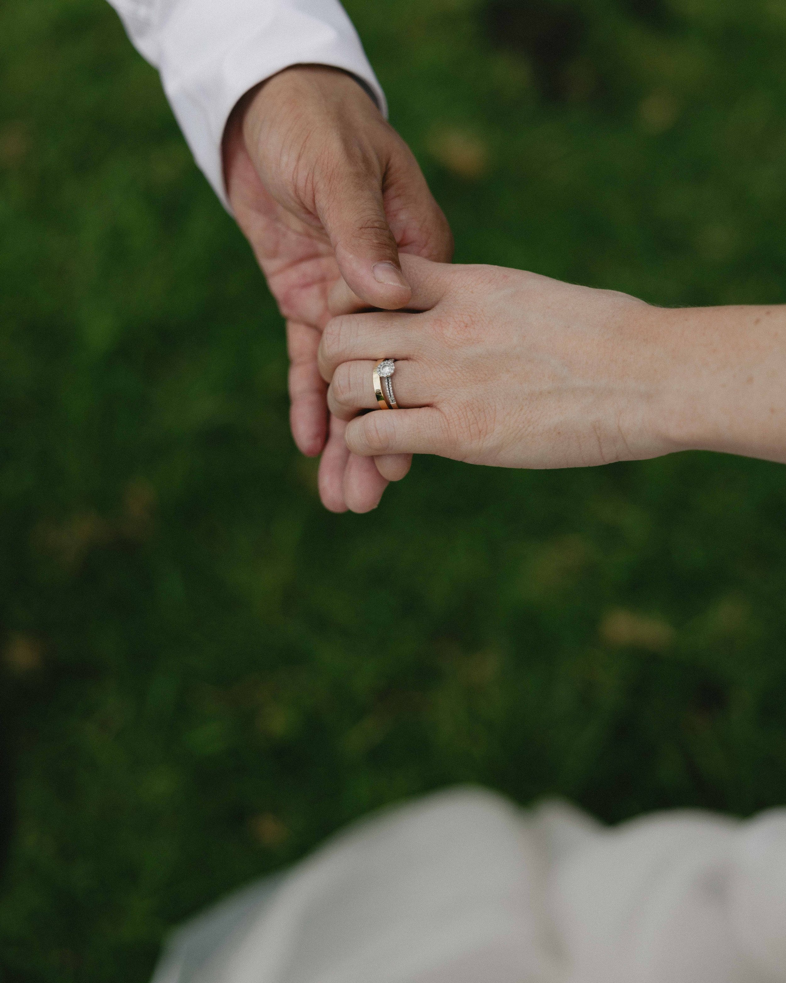 Close-up of two hands holding each other, one with a wedding ring, against a green grassy background.