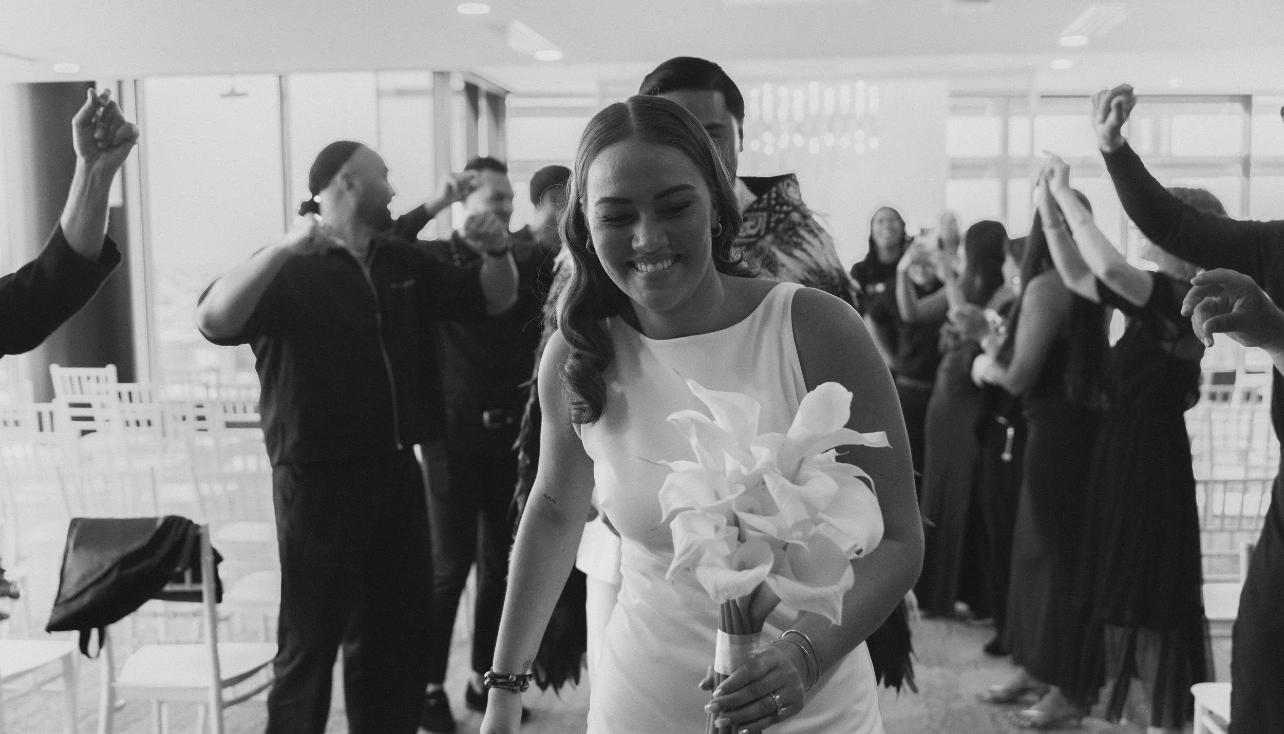 Black and white photo of a woman in a white dress holding a bouquet of flowers, smiling, surrounded by people celebrating and dancing at an indoor event.