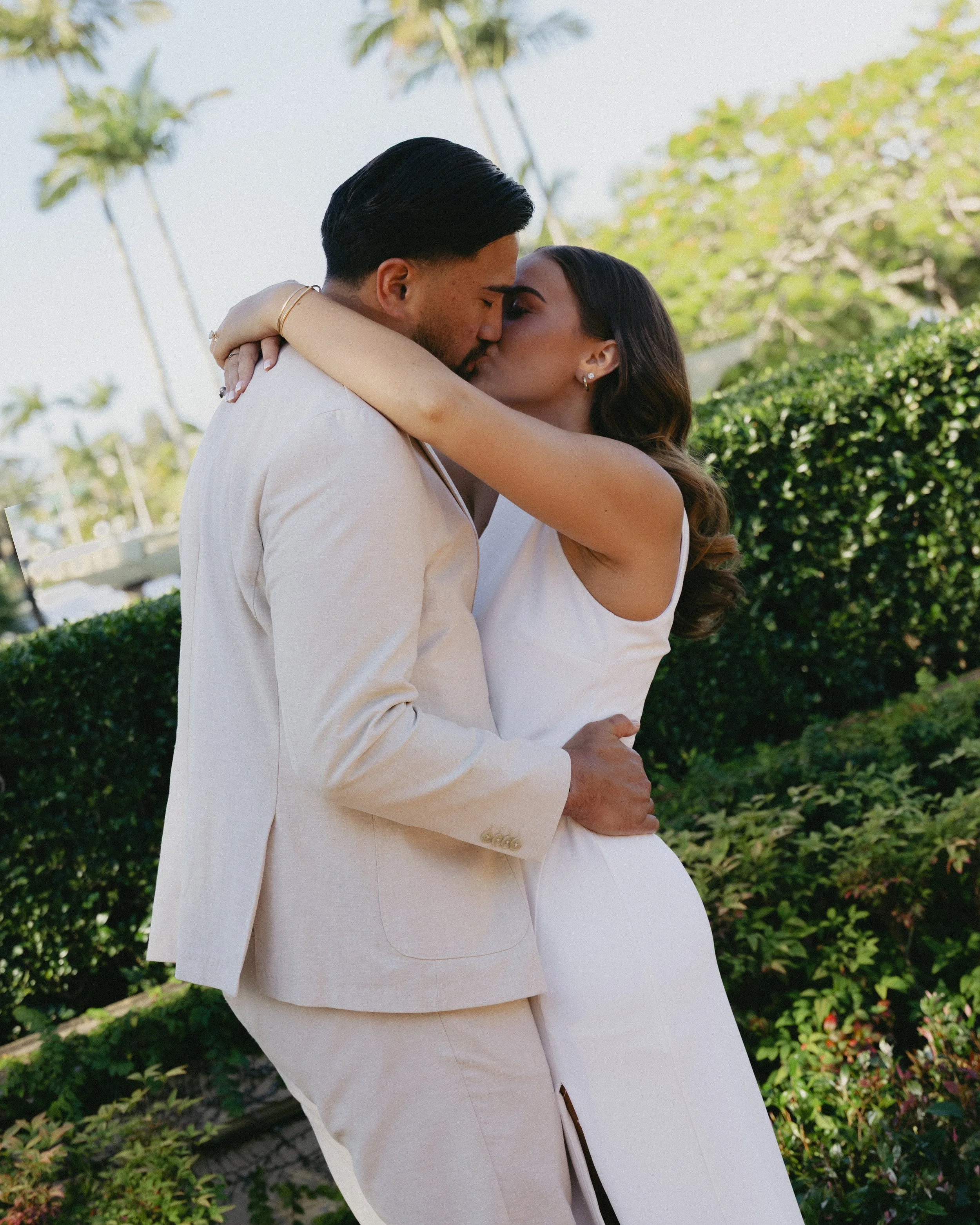 A couple dressed in white kissing outdoors surrounded by greenery and palm trees