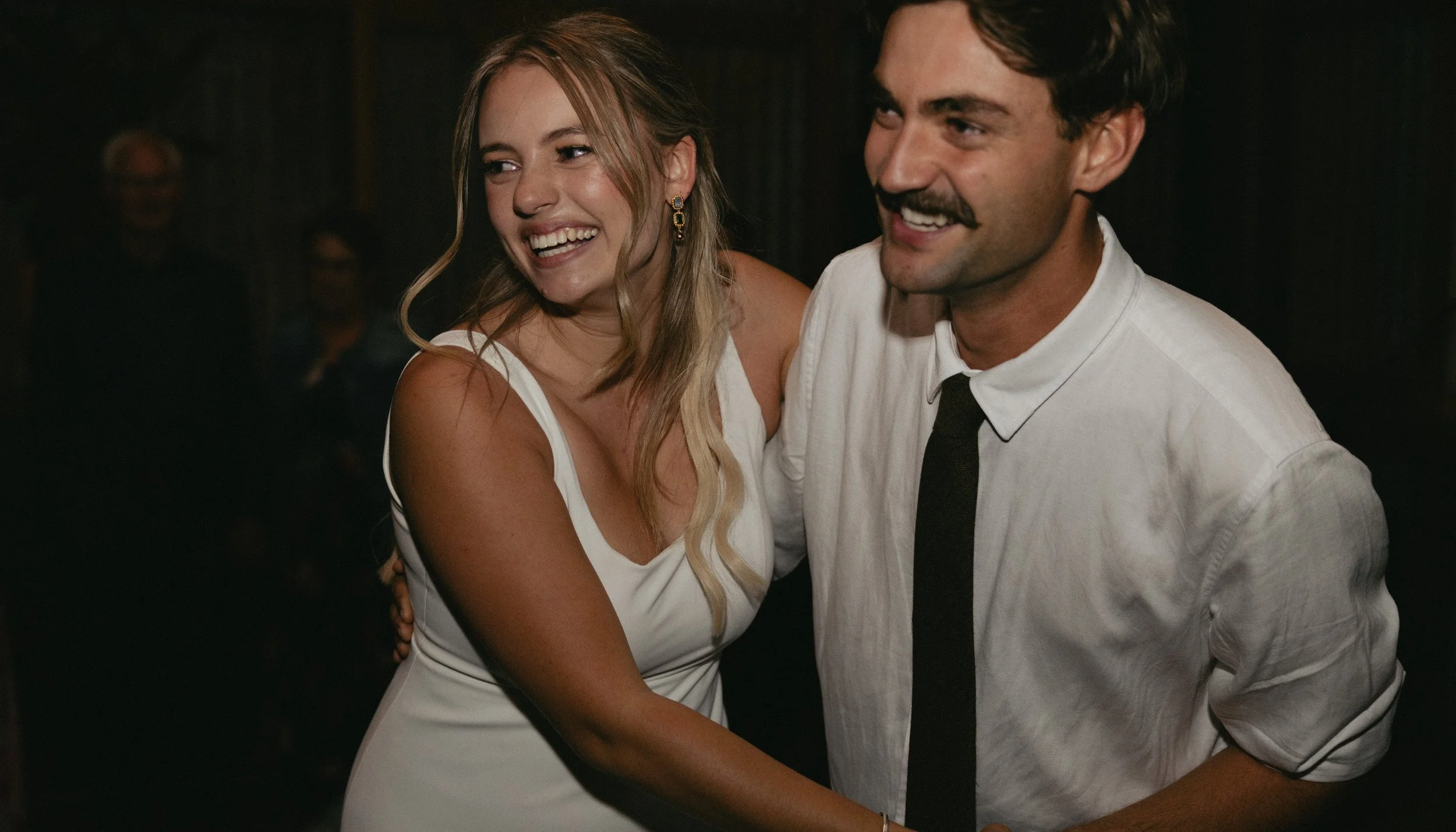 A man and woman dancing and smiling at an event, with other people in the background.