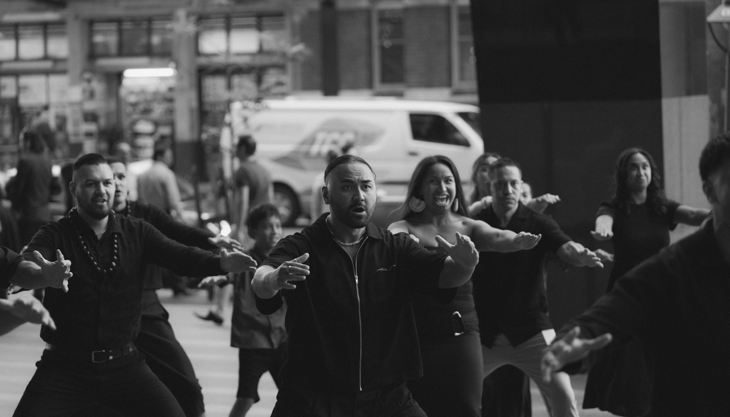A group of people participating in a dance or fitness class in an outdoor urban setting, with some people raising their arms and showing expressions of focus and enthusiasm, in black and white.