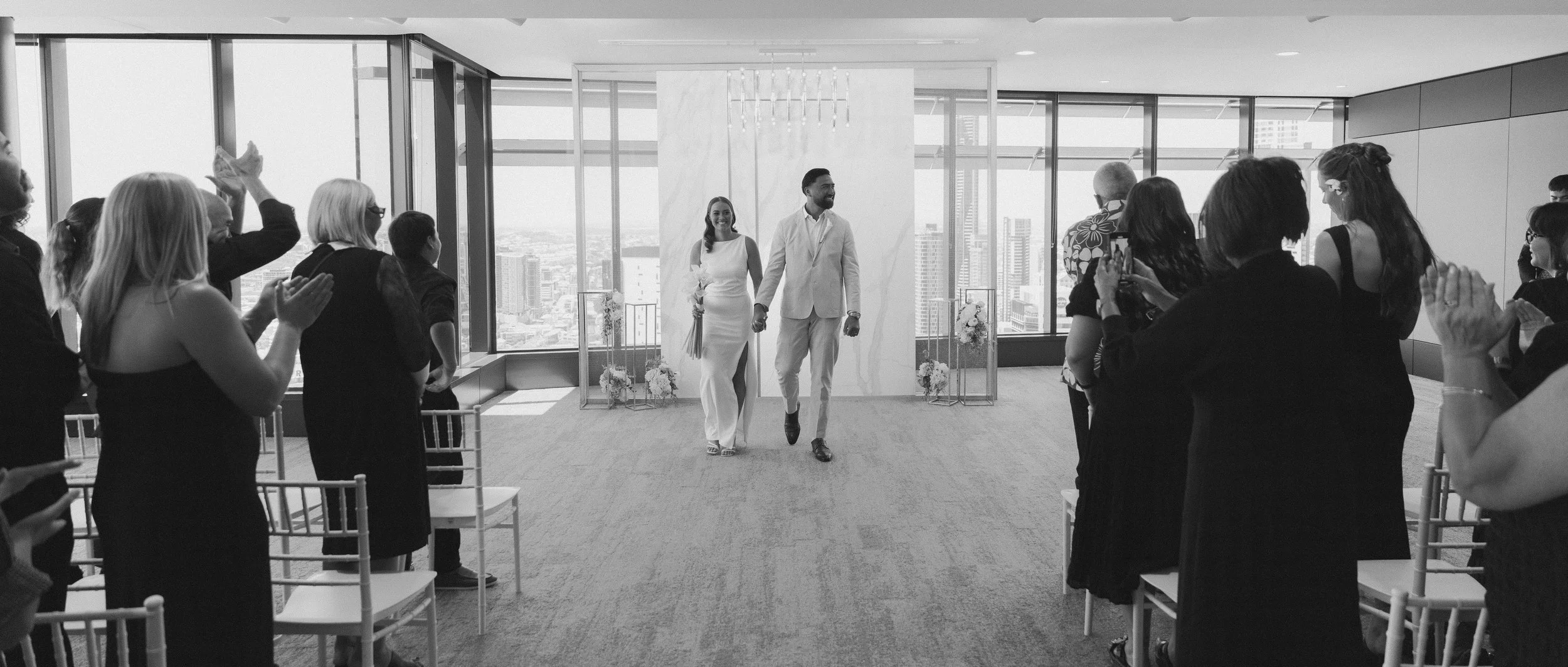 A couple, dressed in white wedding attire, holding hands and walking down the aisle of a modern, high-rise wedding venue with large windows and a city skyline view, while guests stand and applaud.