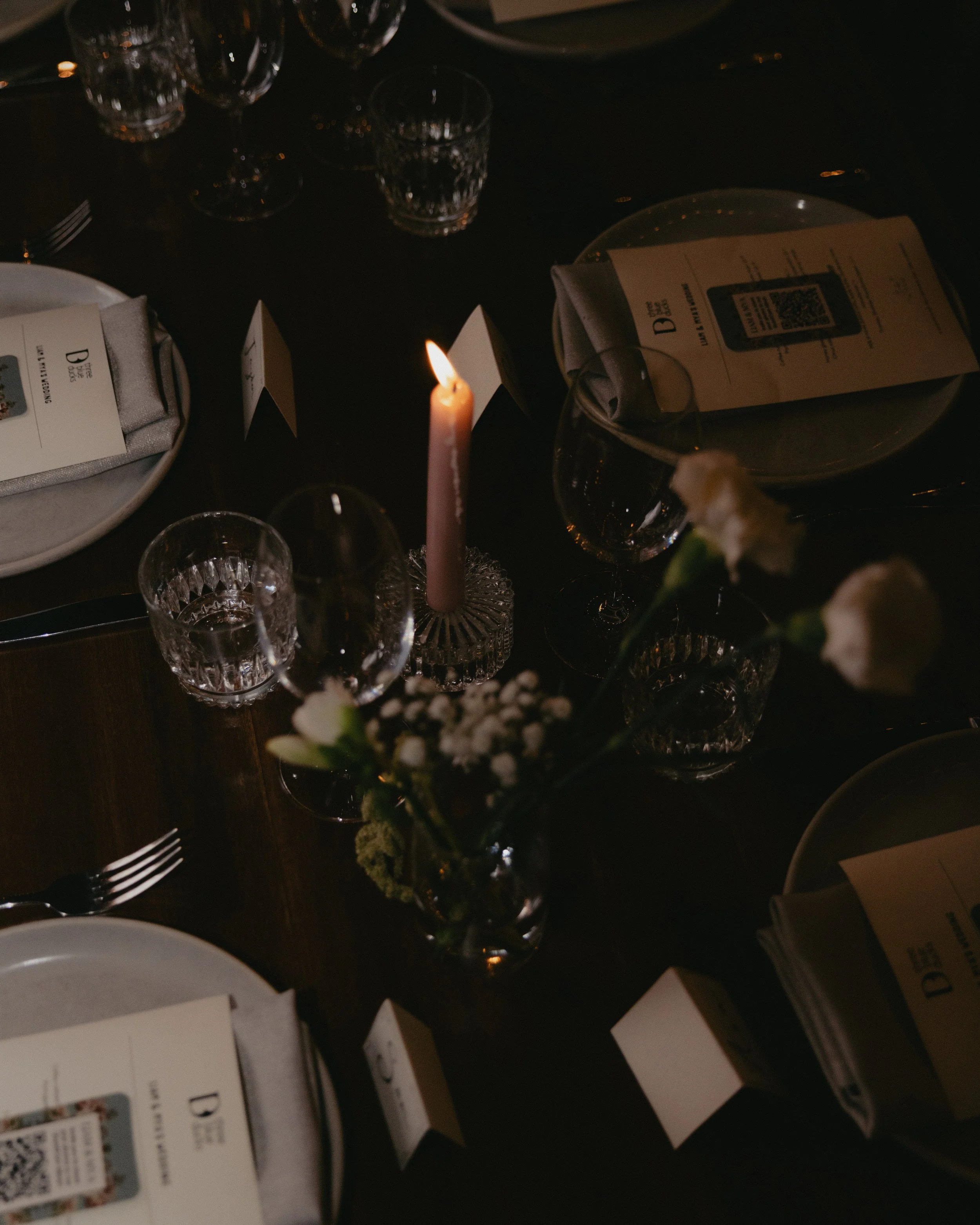 A dark table setting with a lit pink candle, flower vase, wine glasses, plates, silverware, napkins, and small table numbers, illuminated by soft lighting.