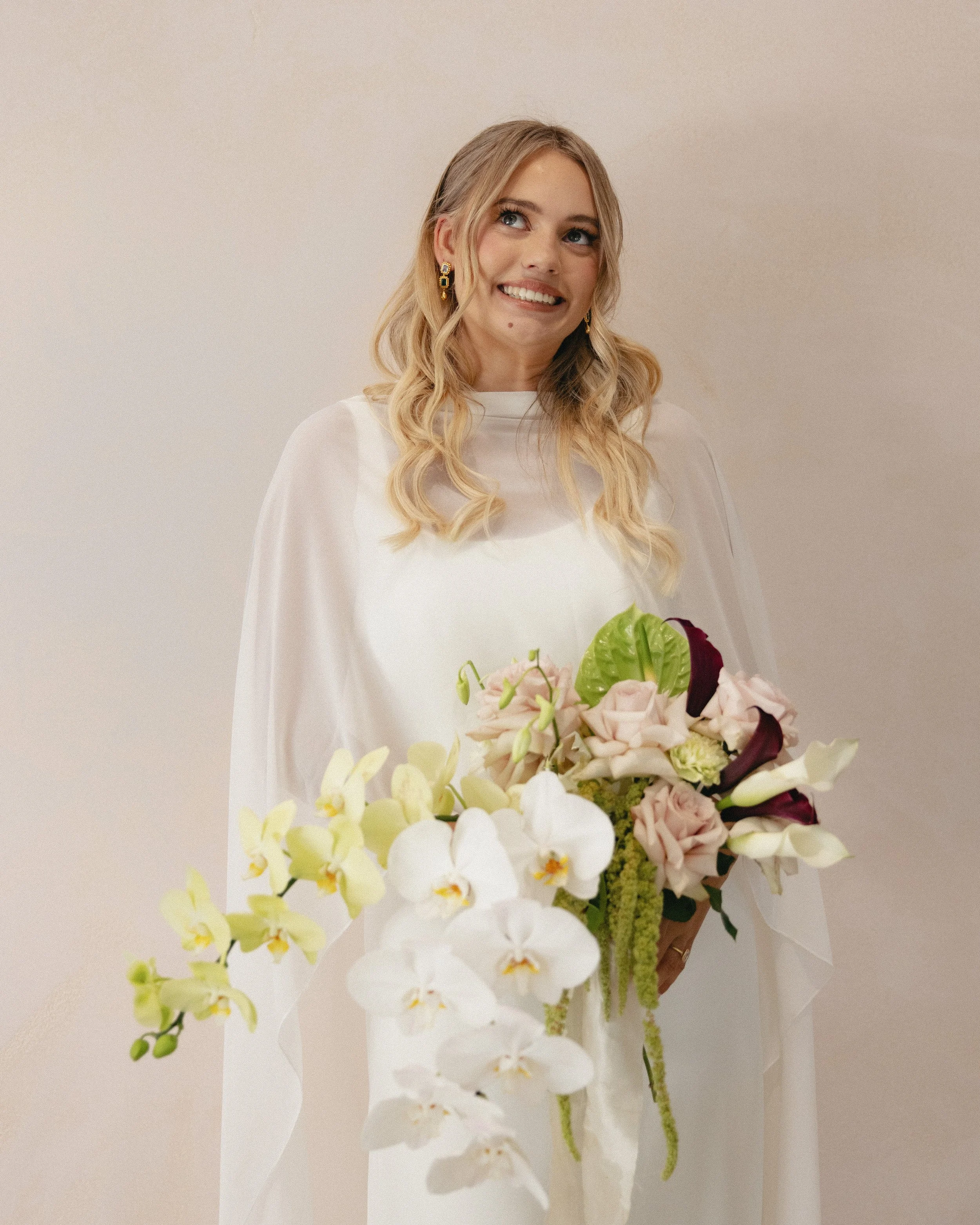 Woman in a white dress holding a large bouquet of flowers, smiling, against a plain background.