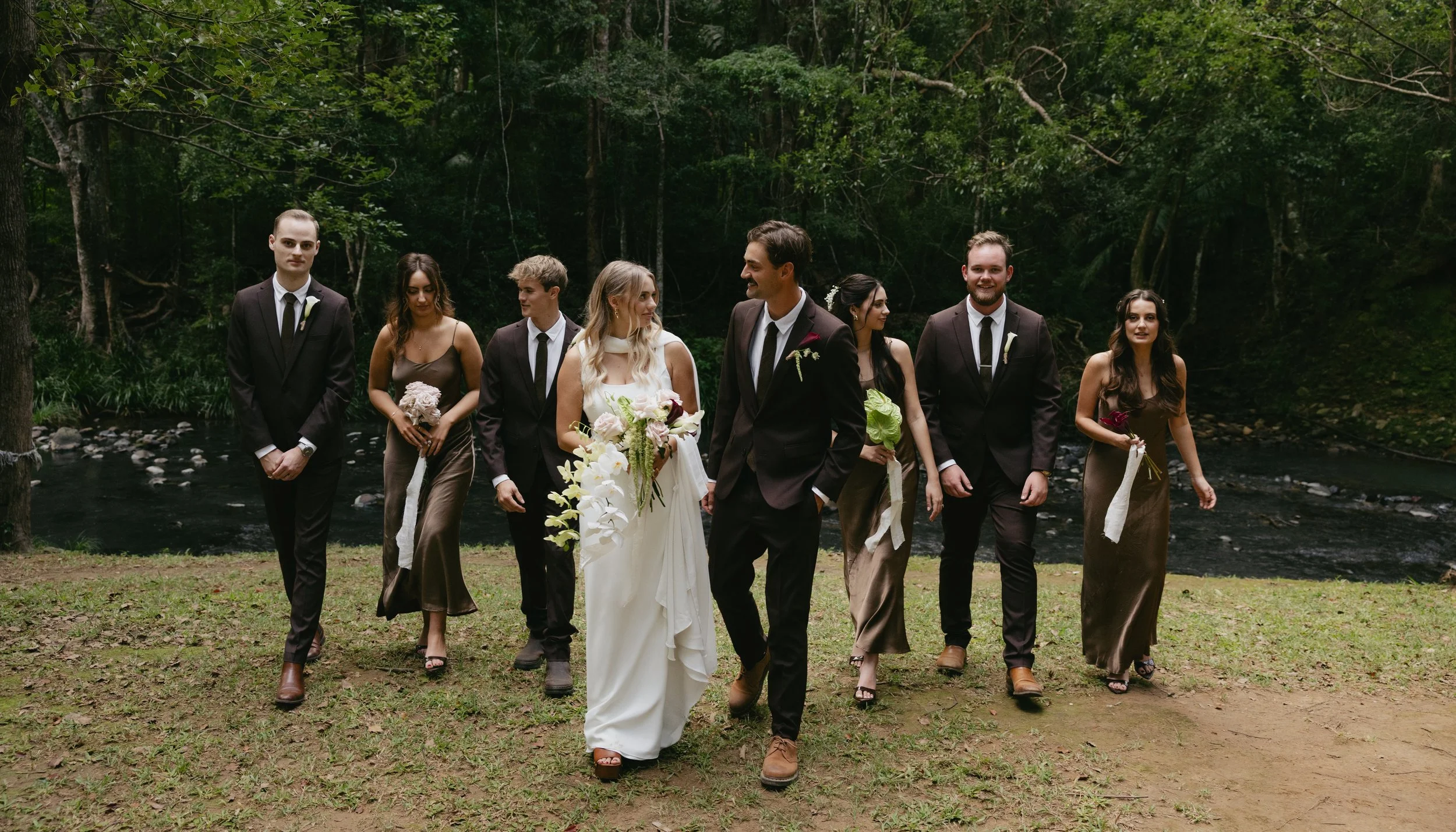 A wedding party walking outdoors near a river in a forest, with the bride in a white dress holding a bouquet, the groom in a black suit, and six bridesmaids and groomsmen in formal attire.
