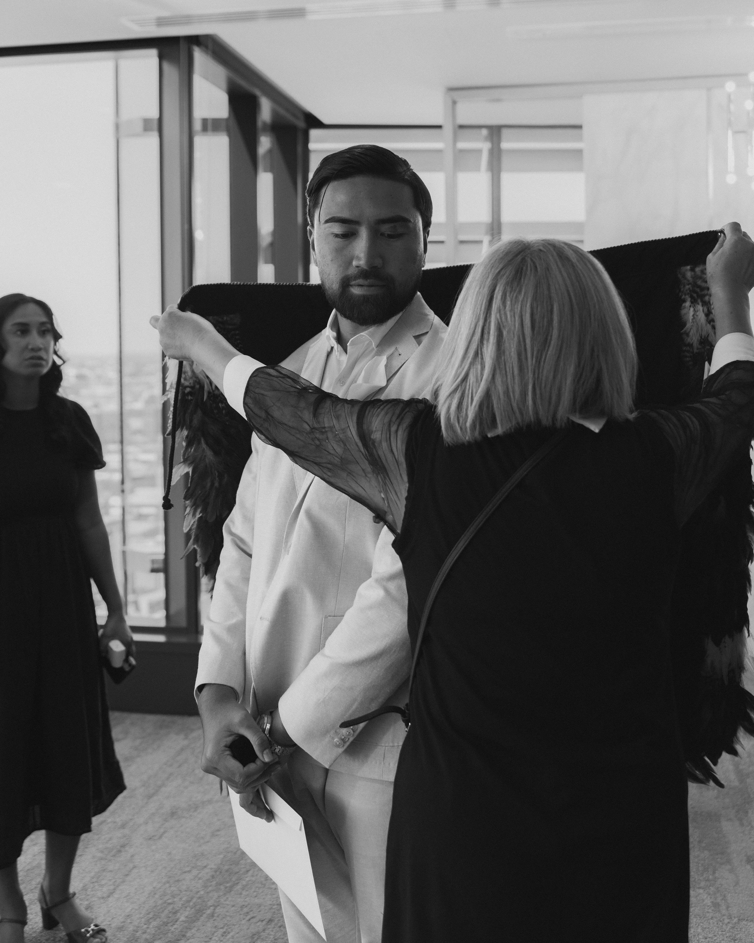 A woman with angel wings is placing a black cape or shawl over a man in a white suit, while another woman observes in the background.