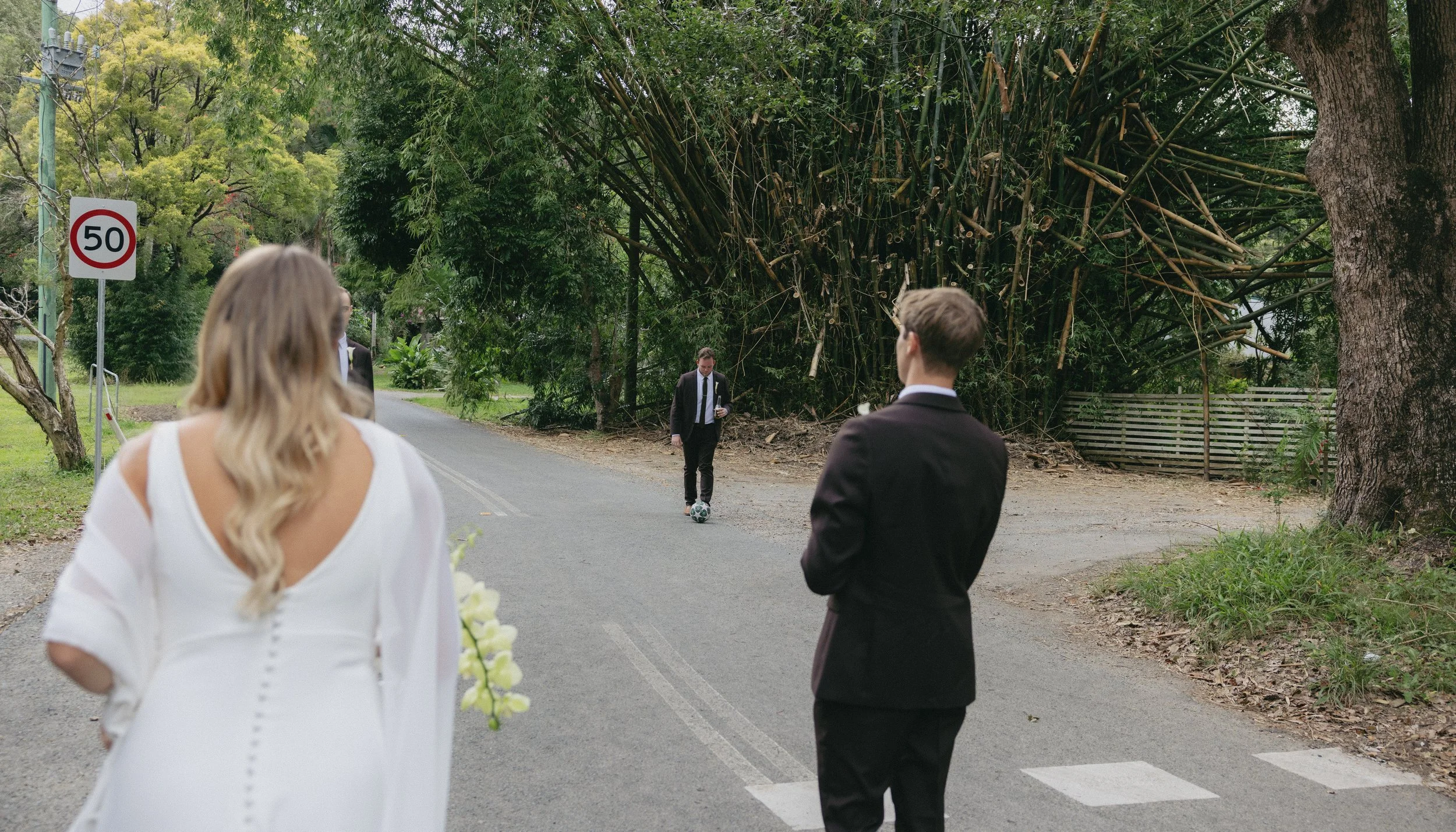 A woman in a white dress holding a bouquet waits on the side of a street, as two men in formal suits, one of whom is kicking a soccer ball, stand nearby. The scene is outdoors with trees and a 50 km/h speed limit sign.