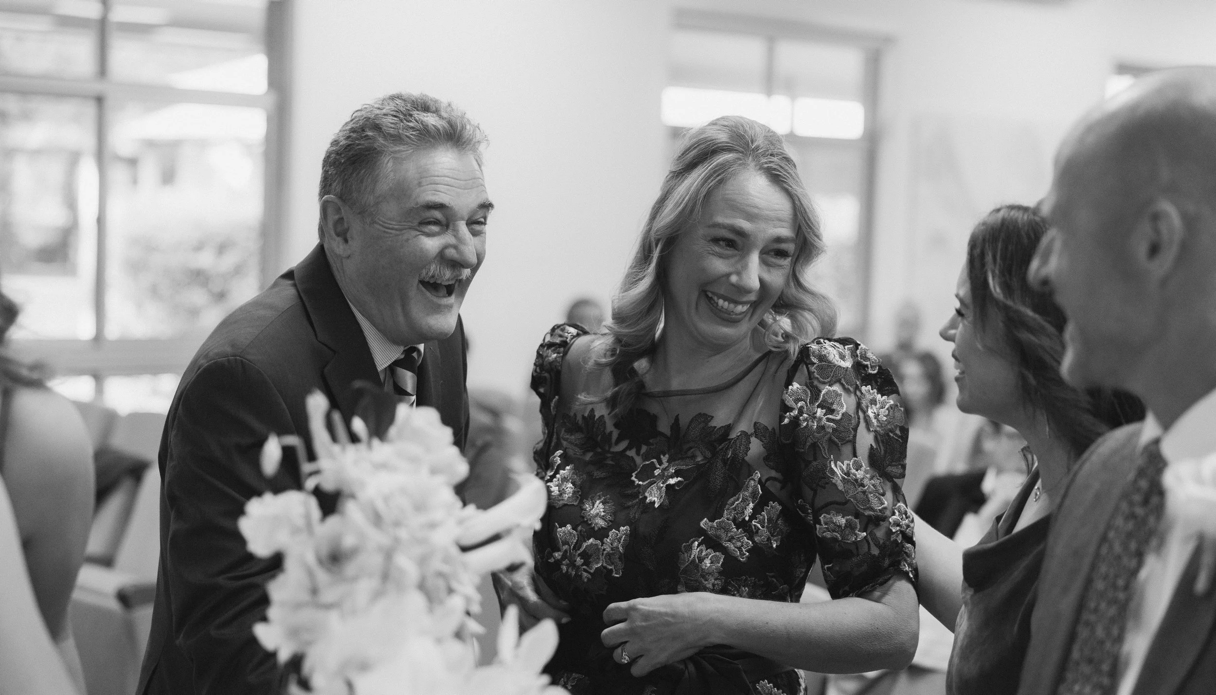 A group of people, including a man with a mustache and a woman with blonde hair, laughing and smiling at a social gathering indoors.