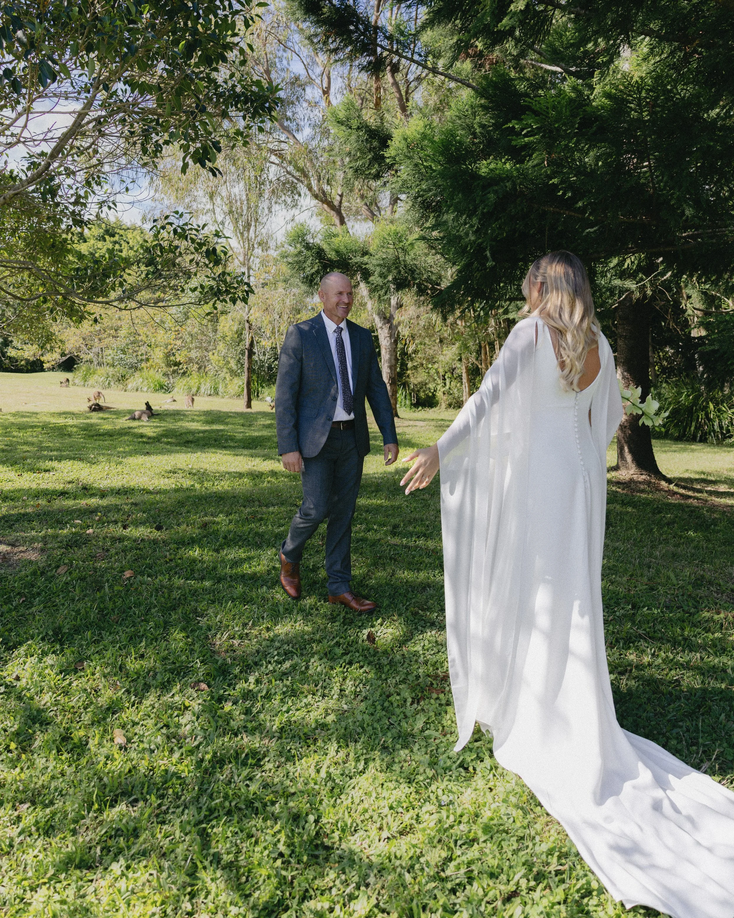 A bride and groom greeting each other outdoors on a sunny day. The bride wears a white wedding dress with long sleeves and a train, while the groom is dressed in a dark suit and tie. They are standing on a grassy area surrounded by trees.
