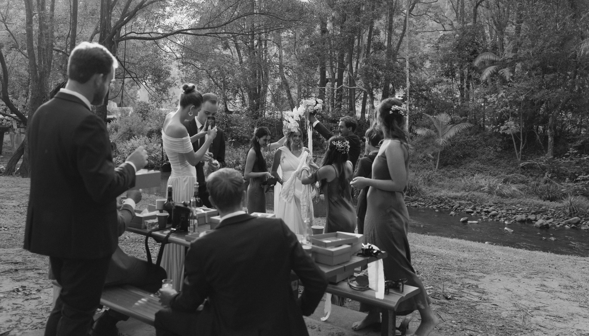 A group of people at an outdoor wedding celebration near a river, some taking photos, others dancing, with trees and nature in the background, in black and white.