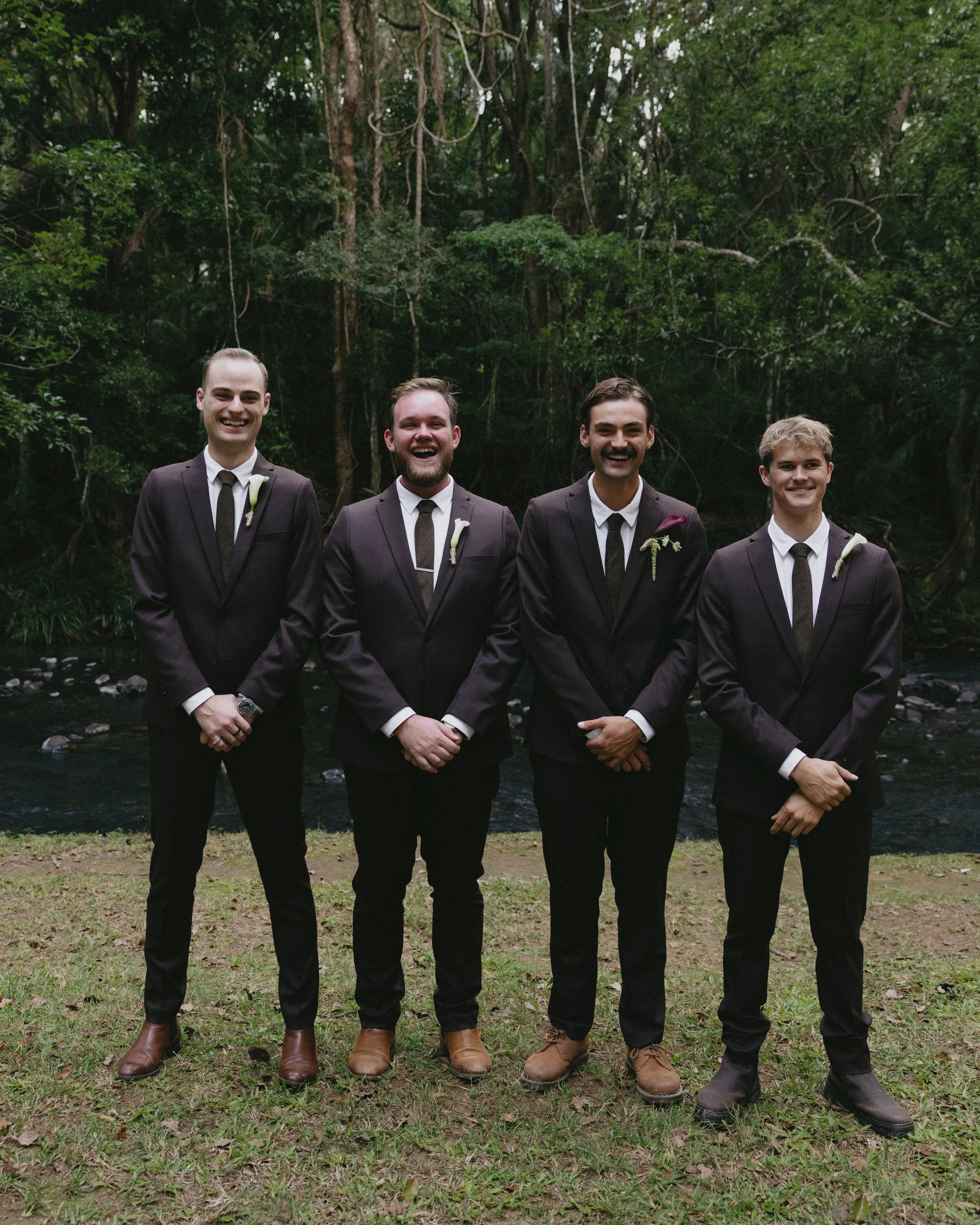 Four men dressed in dark suits and ties standing outdoors in front of a forest with a small stream, smiling for the camera.