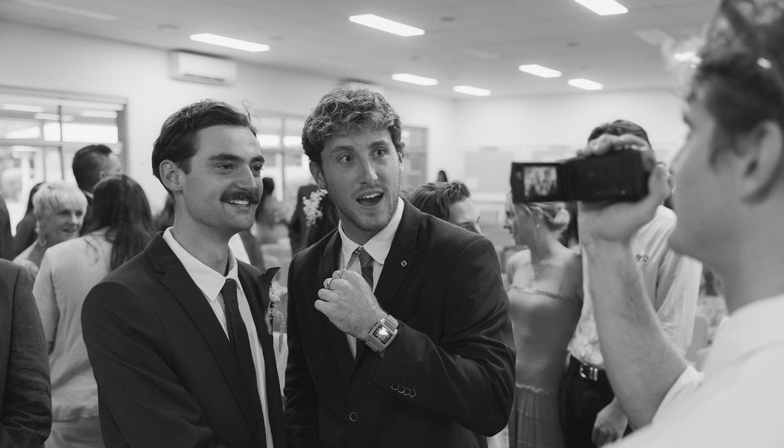 Two men in suits smiling and talking to each other at a social gathering with other people in the background, some smiling and some engaging in different activities, in a bright indoor space.