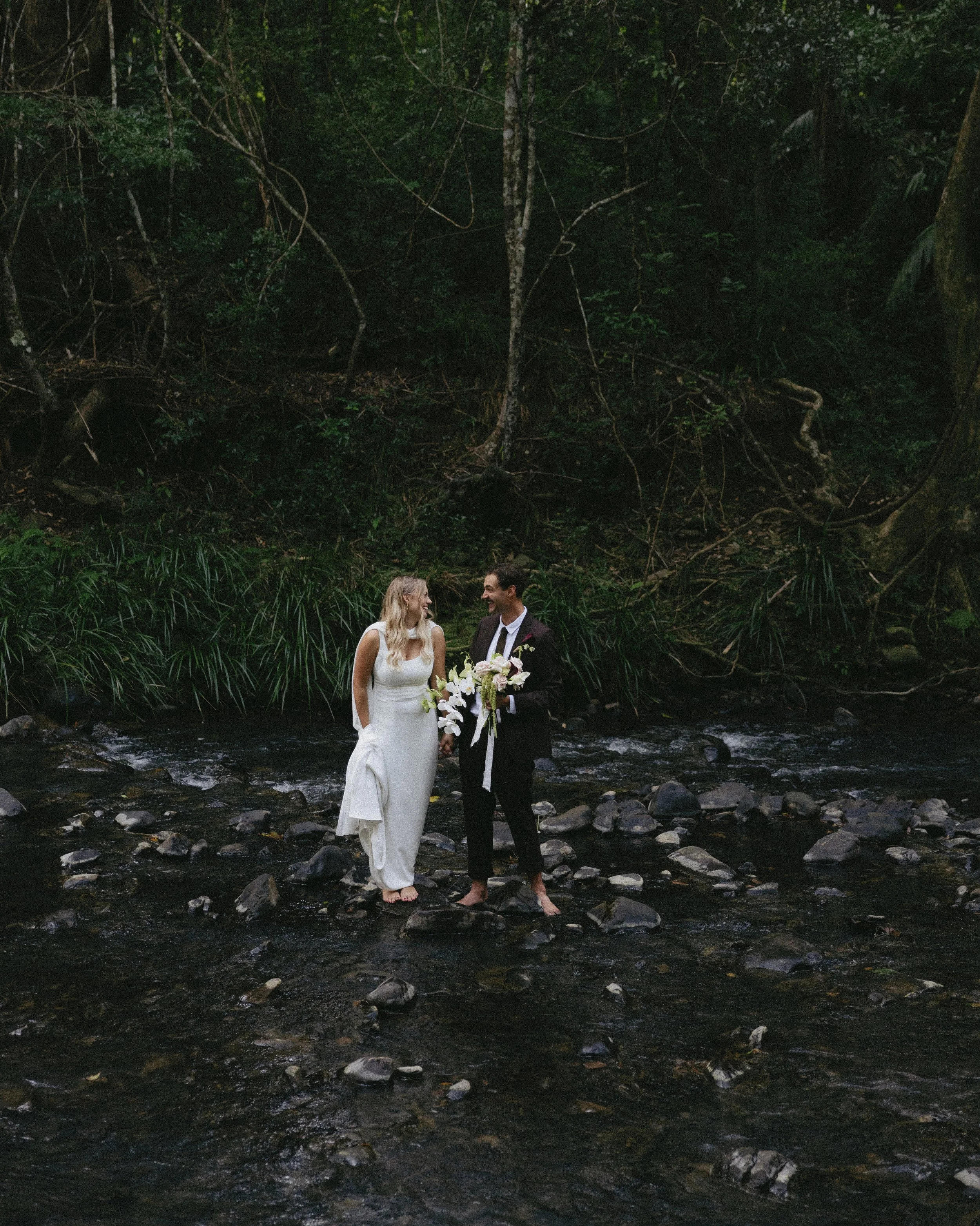 A couple dressed in wedding attire is standing on rocks in a shallow stream, holding hands and smiling at each other with a lush green forest in the background.