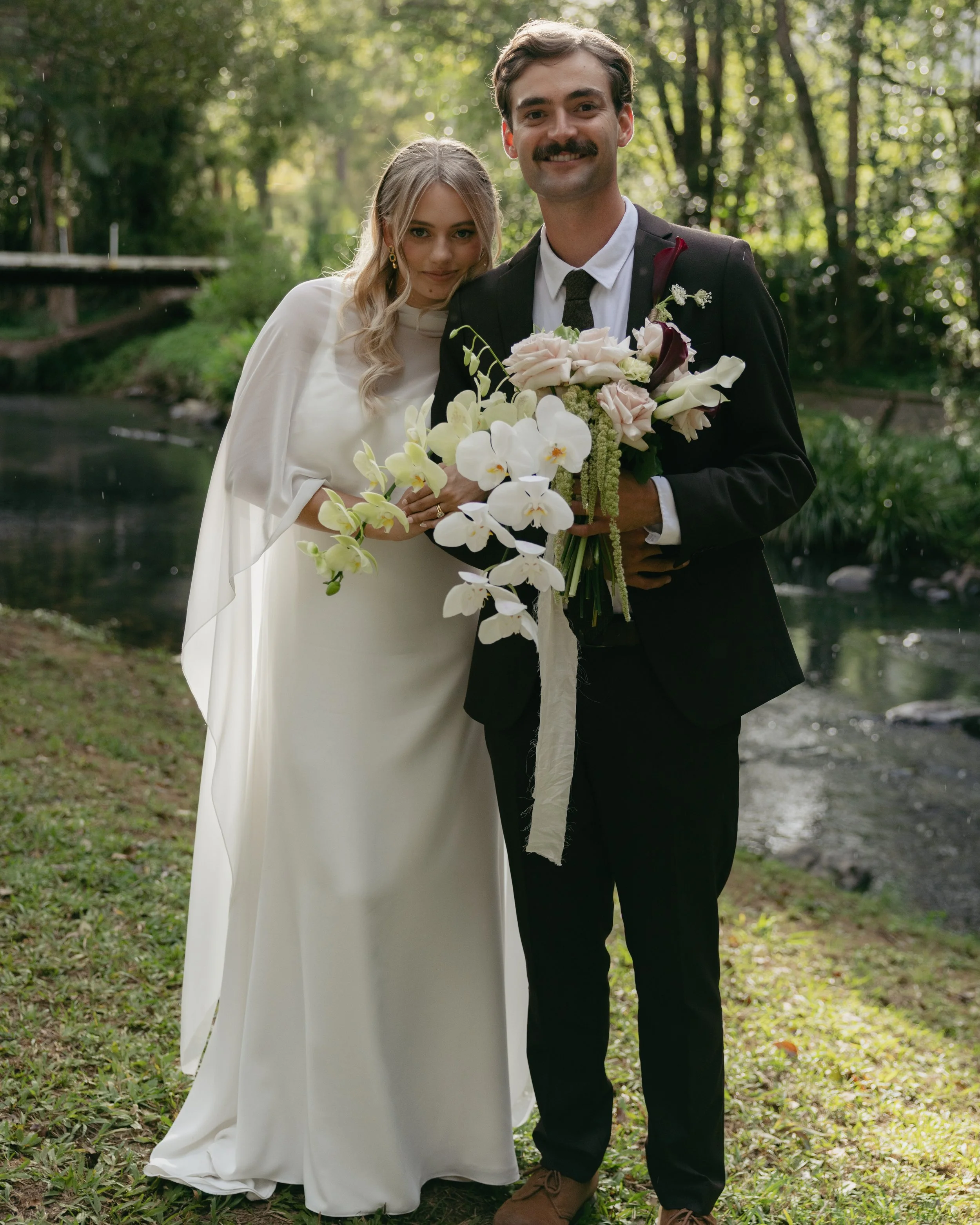 A bride and groom standing together outdoors near a stream, dressed in wedding attire, holding a bouquet of white and blush flowers, with trees and greenery in the background.