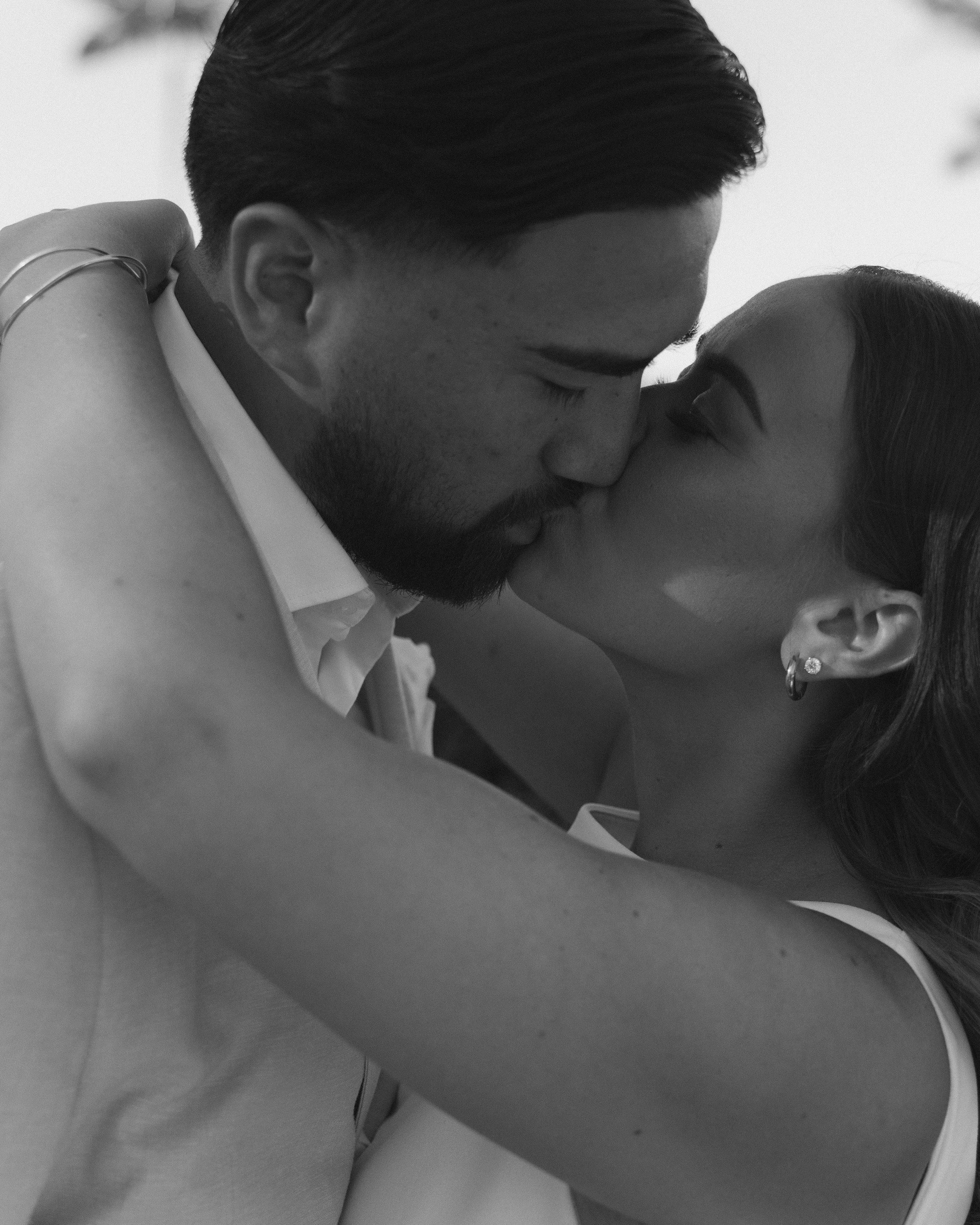 A black and white photo of a couple kissing, showing their faces close together with their eyes closed, and their arms wrapped around each other.