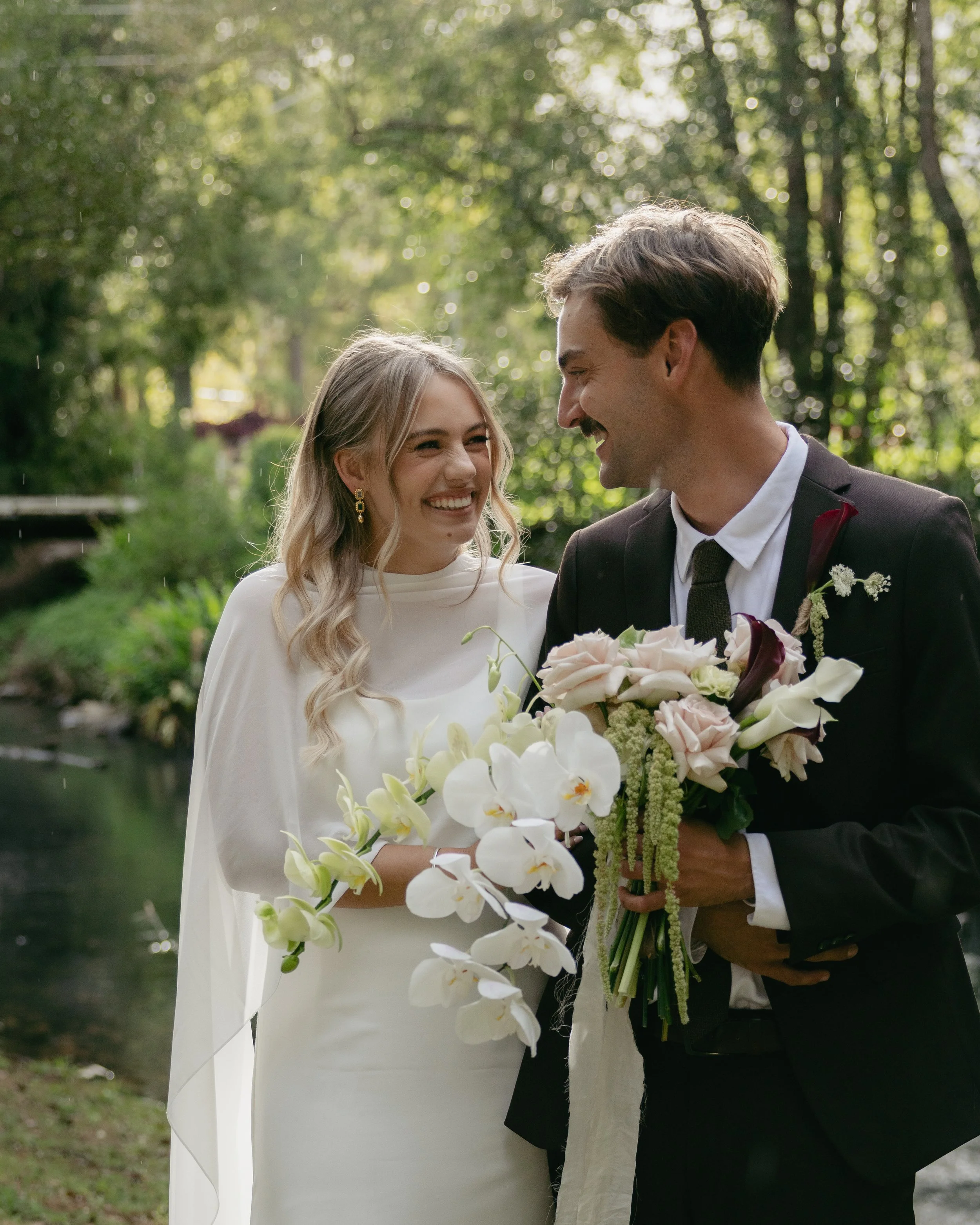 A smiling bride and groom enjoying their wedding outdoors with greenery and a small stream in the background, the groom holding a bouquet of flowers.