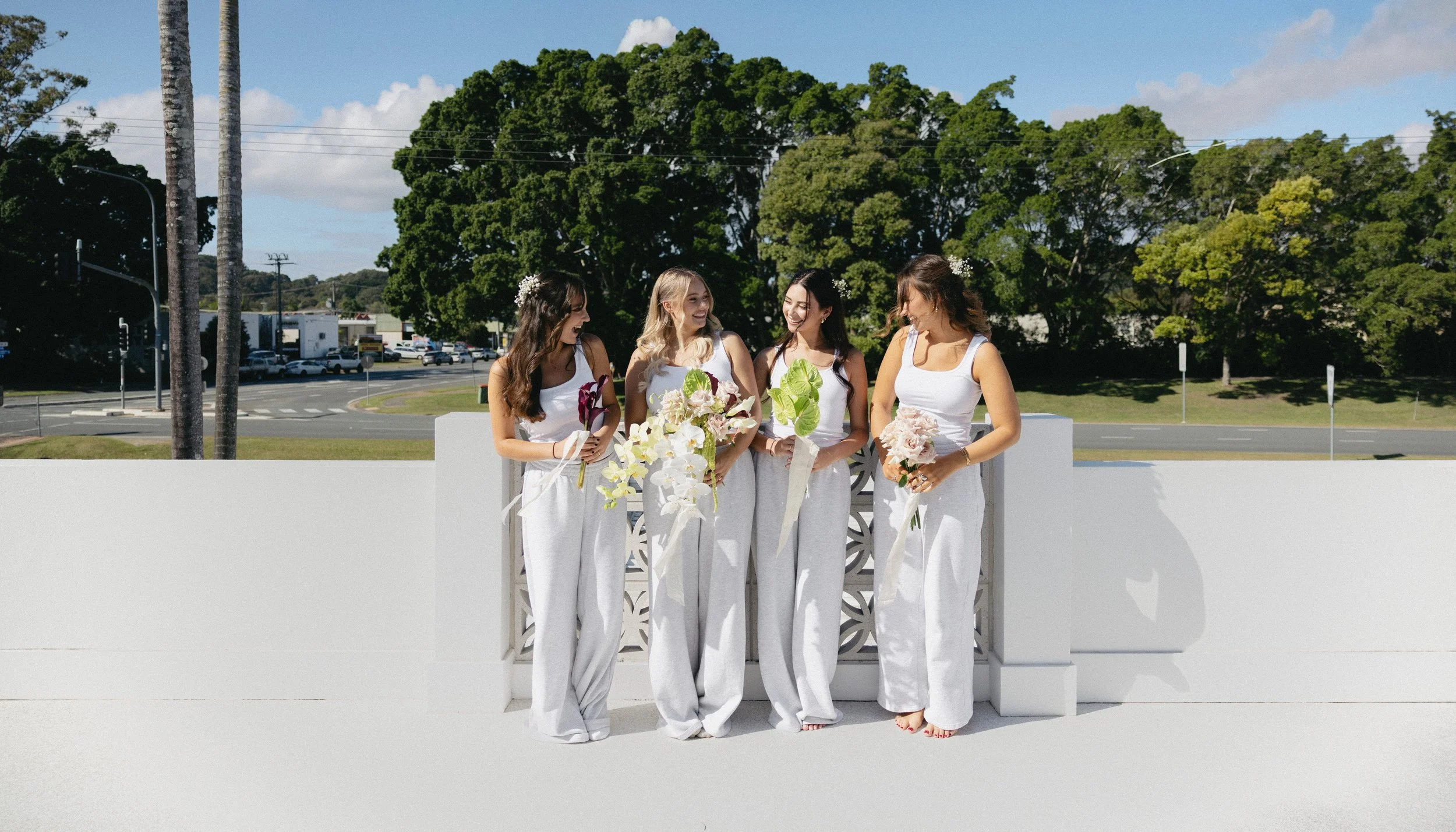 Four women dressed in white and holding floral bouquets, standing outdoors near a white wall with trees and a road in the background, possibly preparing for a wedding or special event.