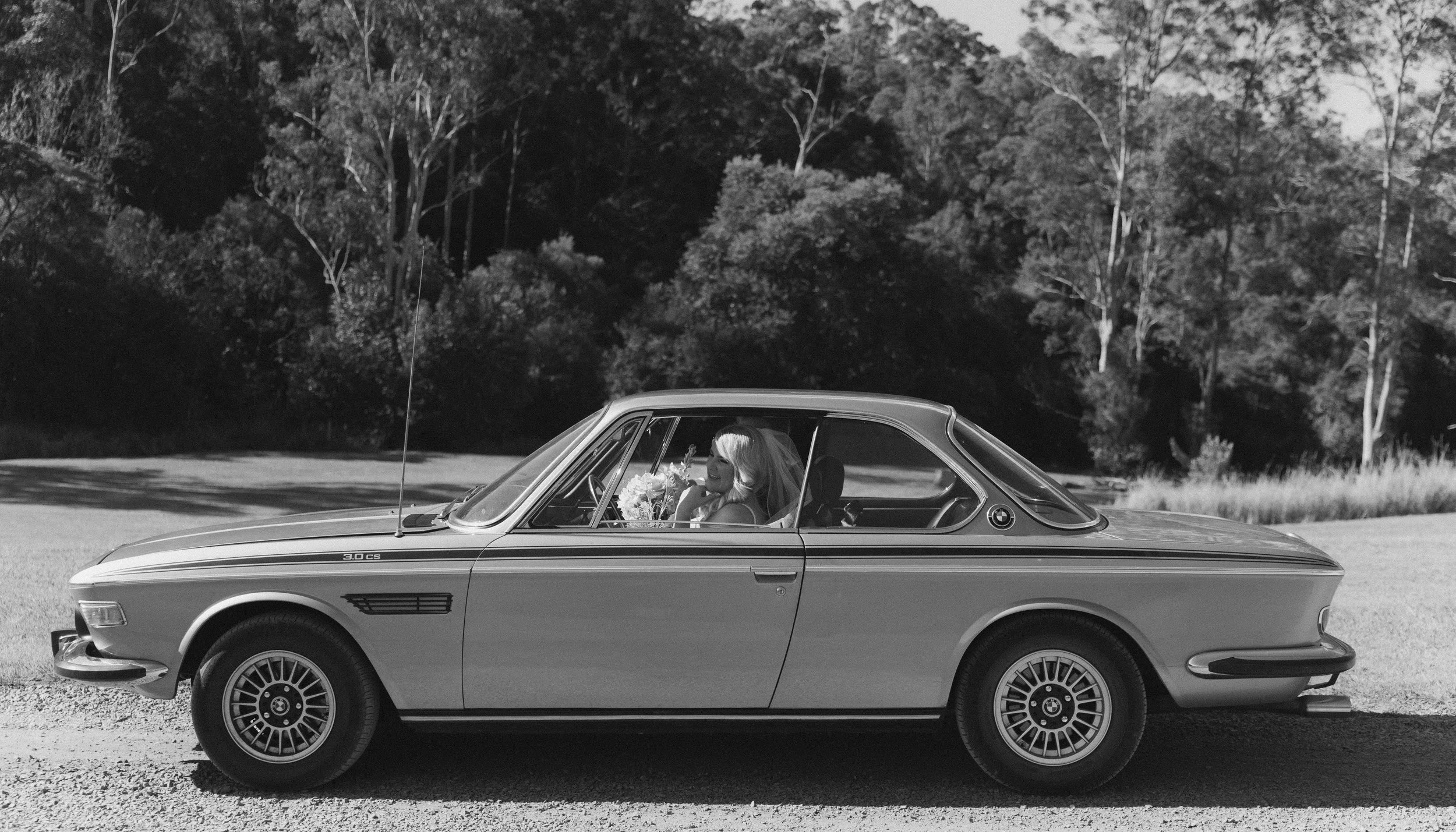 A black and white photo of a woman with blonde hair inside a classic BMW car parked on a gravel road with trees and hills in the background.