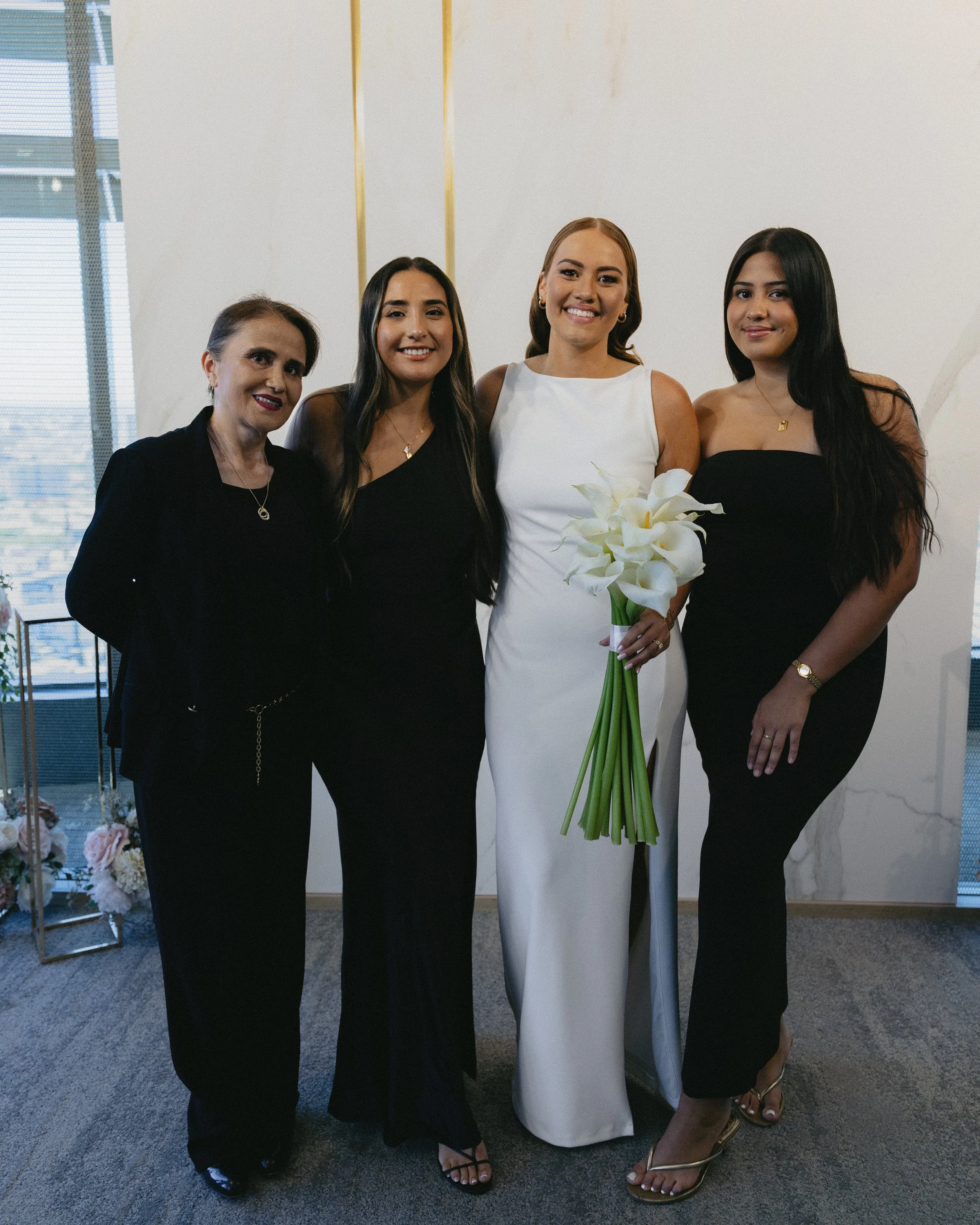 Four women standing together indoors, one of whom is wearing a white dress and holding a bouquet of white calla lilies, likely celebrating a special occasion.
