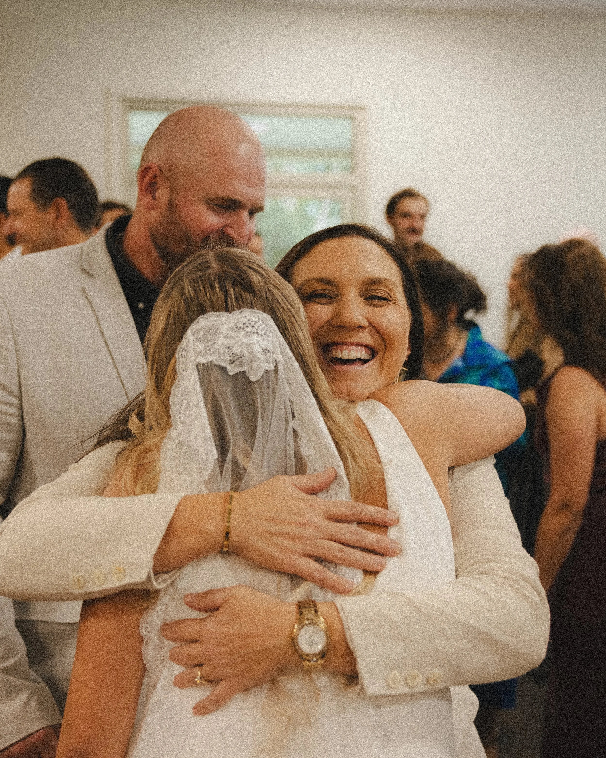 People hugging and smiling at a wedding reception, including a bride wearing a veil and a woman in a white dress hugging her.