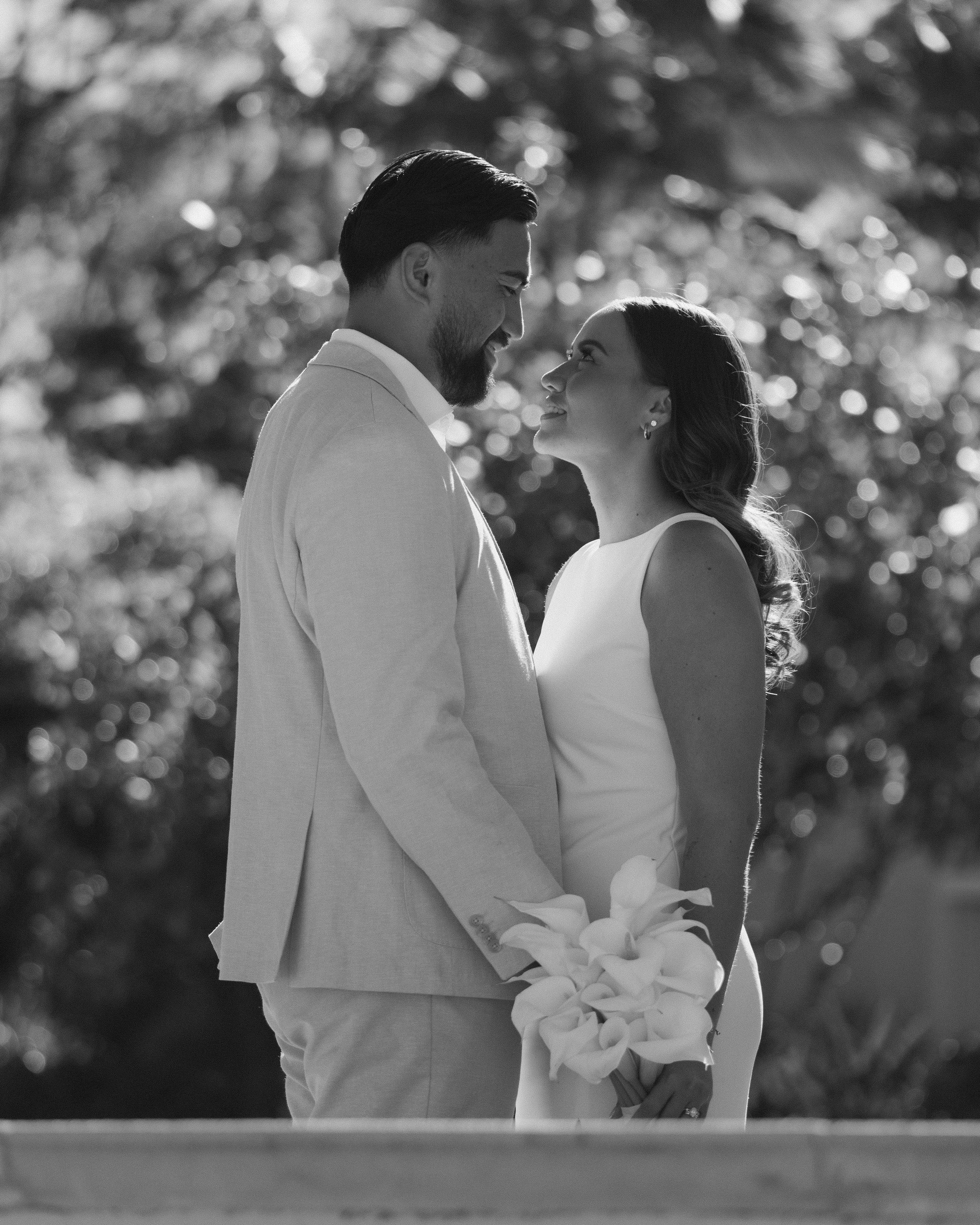 A black-and-white photograph of a bride and groom standing close, holding hands, and gazing into each other's eyes outdoors during daytime. The bride is holding a bouquet of calla lilies.