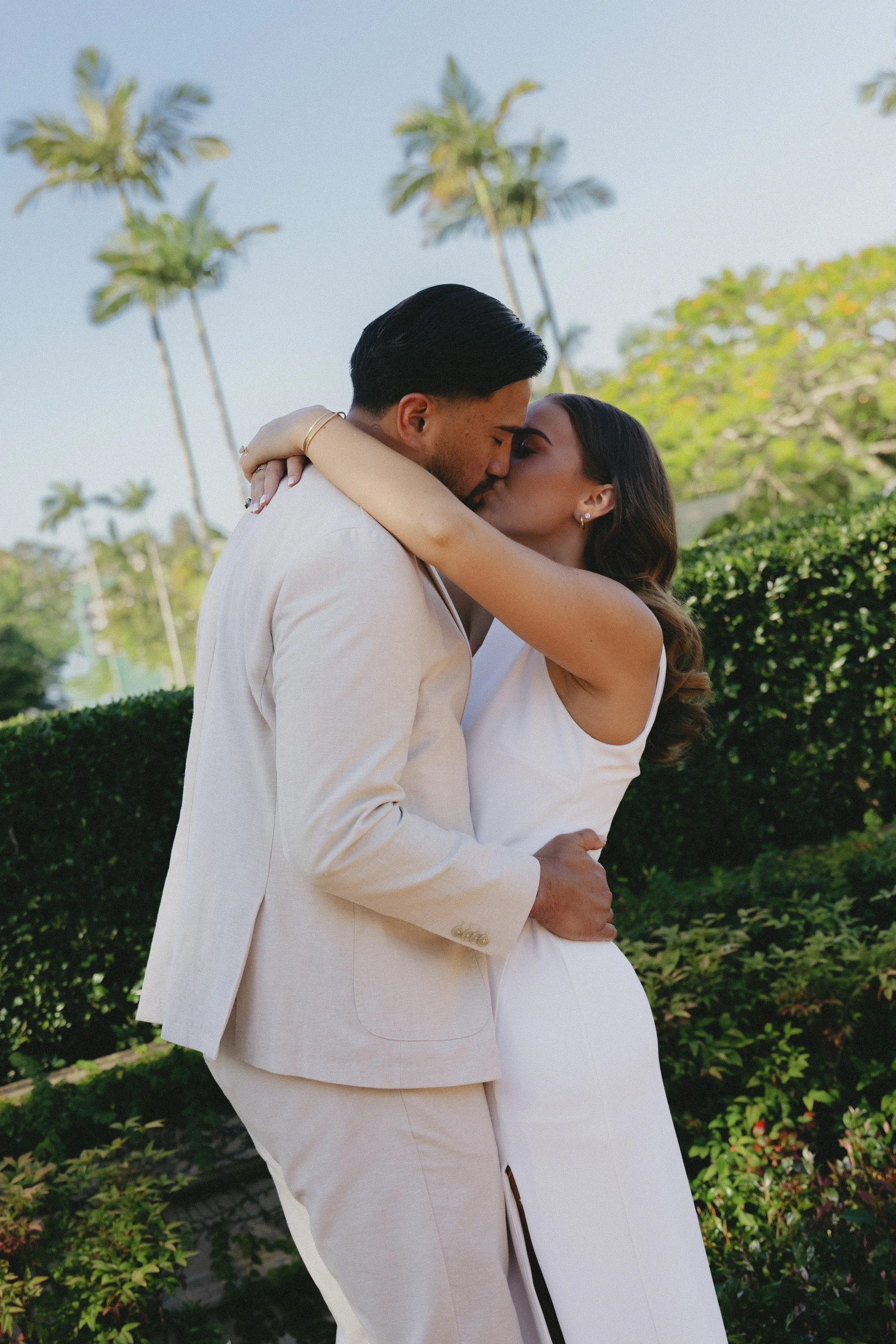 A couple wearing white clothing sharing a kiss outdoors, with palm trees and bushes in the background.